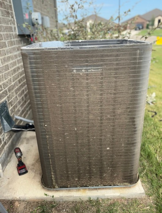 Exterior air conditioning unit, dirty fins, next to a brick wall and green grass.