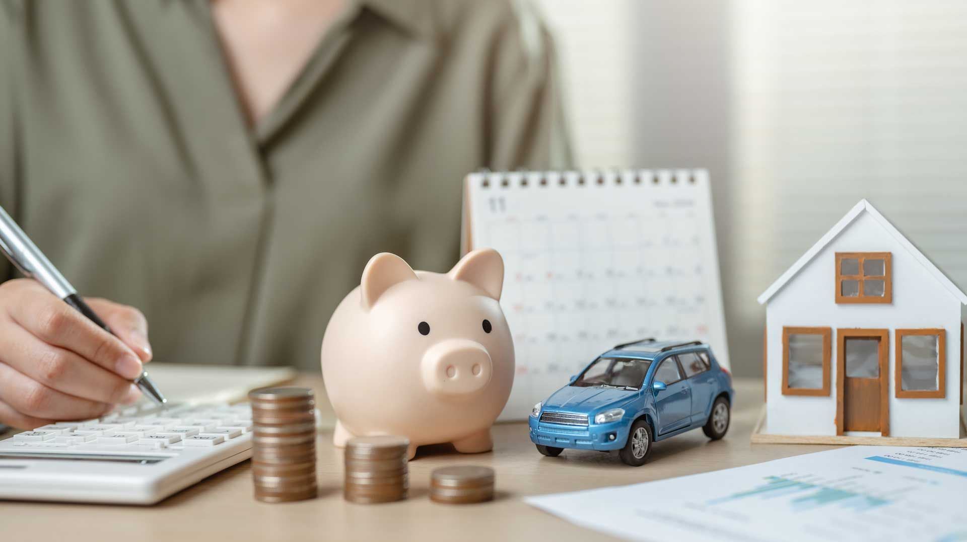 Person calculating finances near a piggy bank, coins, toy car, and house. Person calculating finances near a piggy bank, coins, toy car, and house.