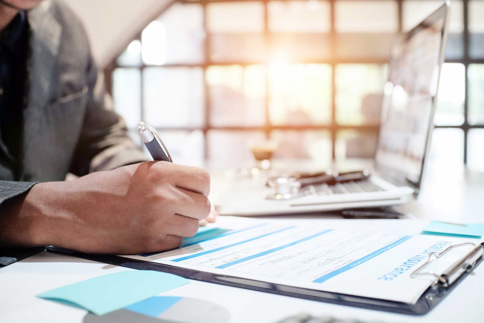 Person writing on a document at a desk with a laptop and bright window in background.