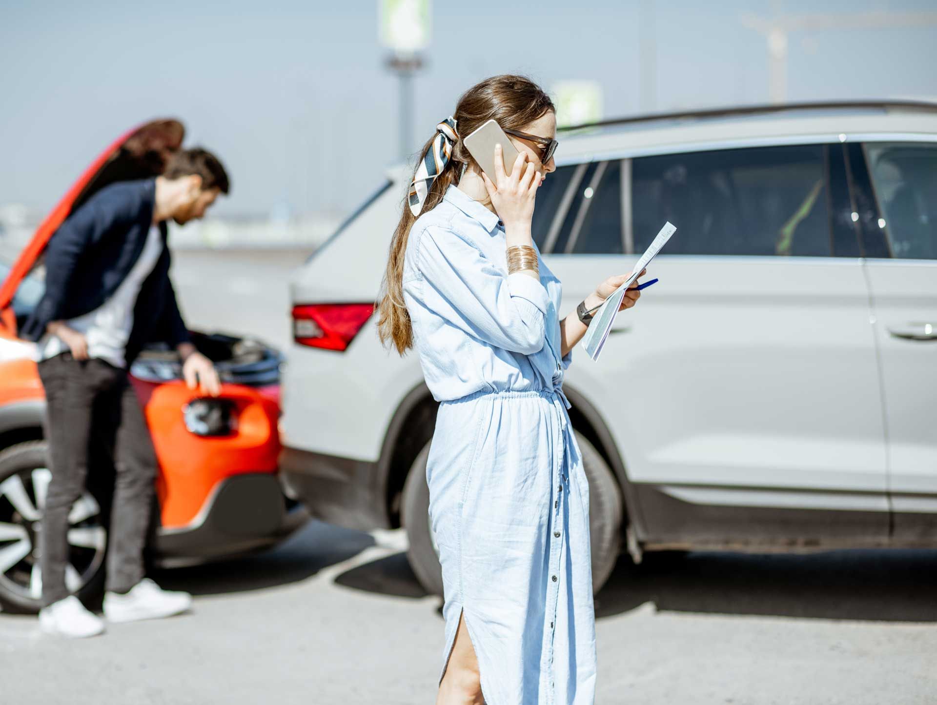 Woman on phone at car accident scene, reviewing paperwork; man inspects damaged orange car.