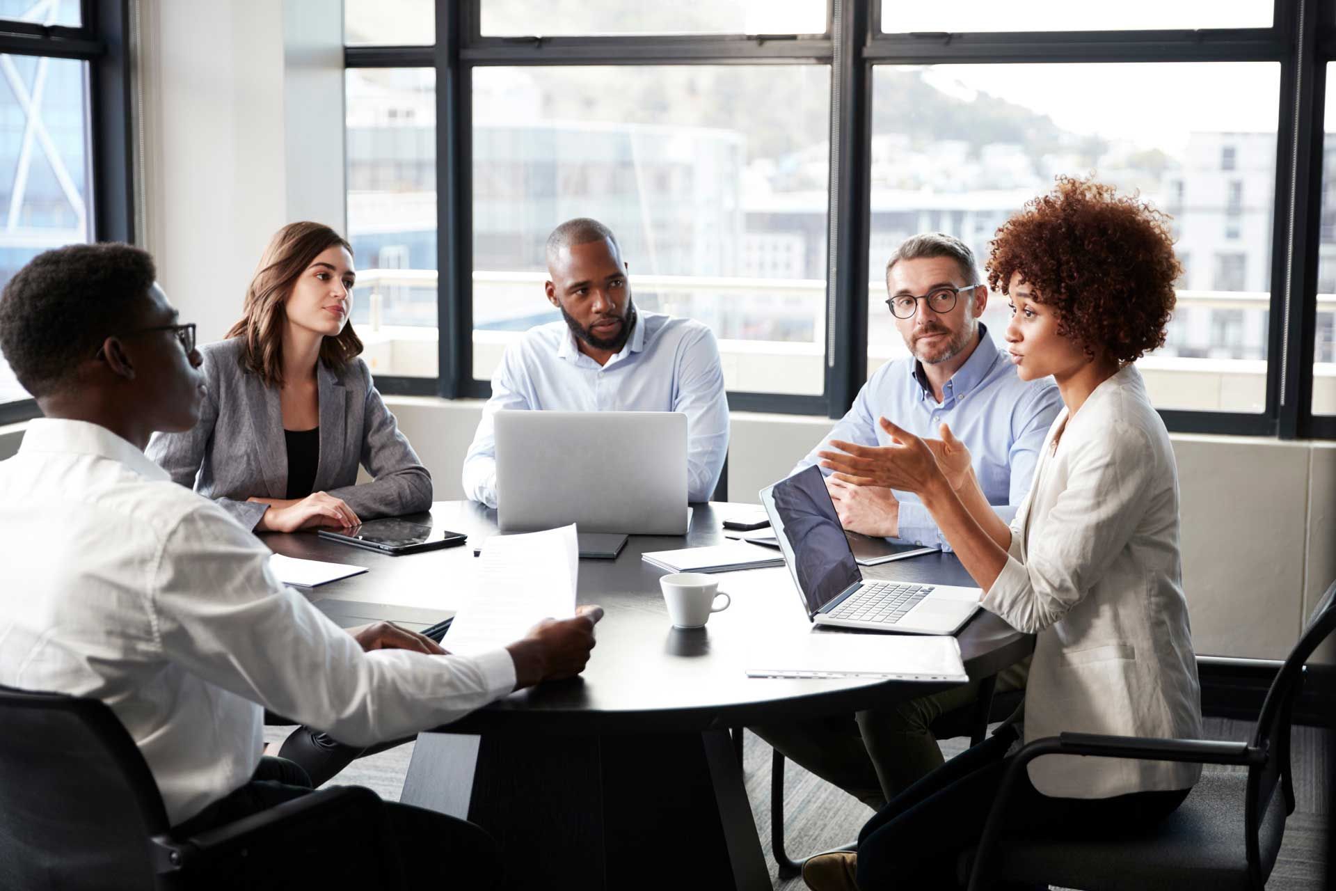 Business meeting in a modern office. People at a round table, discussing with laptops and papers.