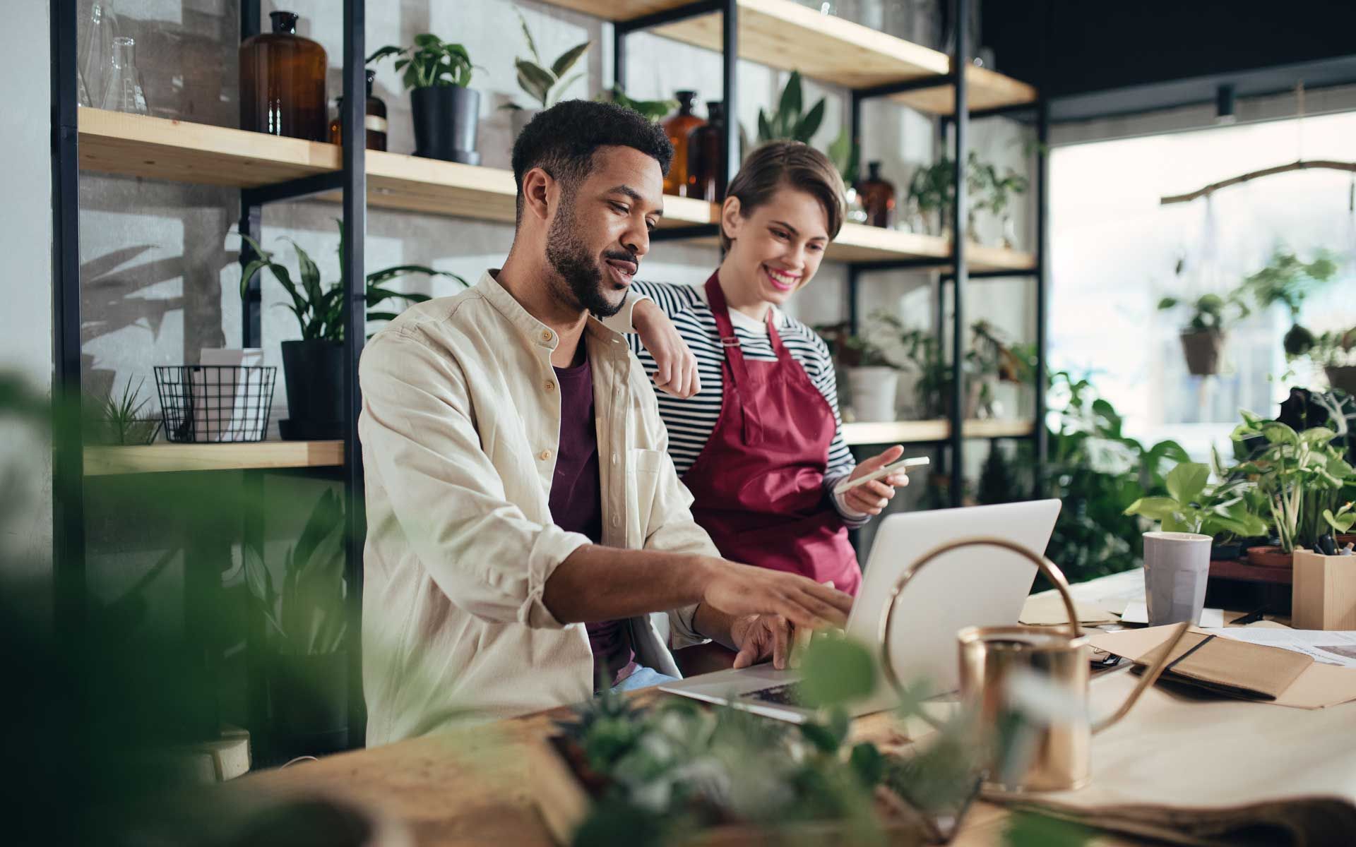 Man and woman working on laptop in a plant shop, surrounded by plants and shelves. Man and woman working on laptop in a plant shop, surrounded by plants and shelves.