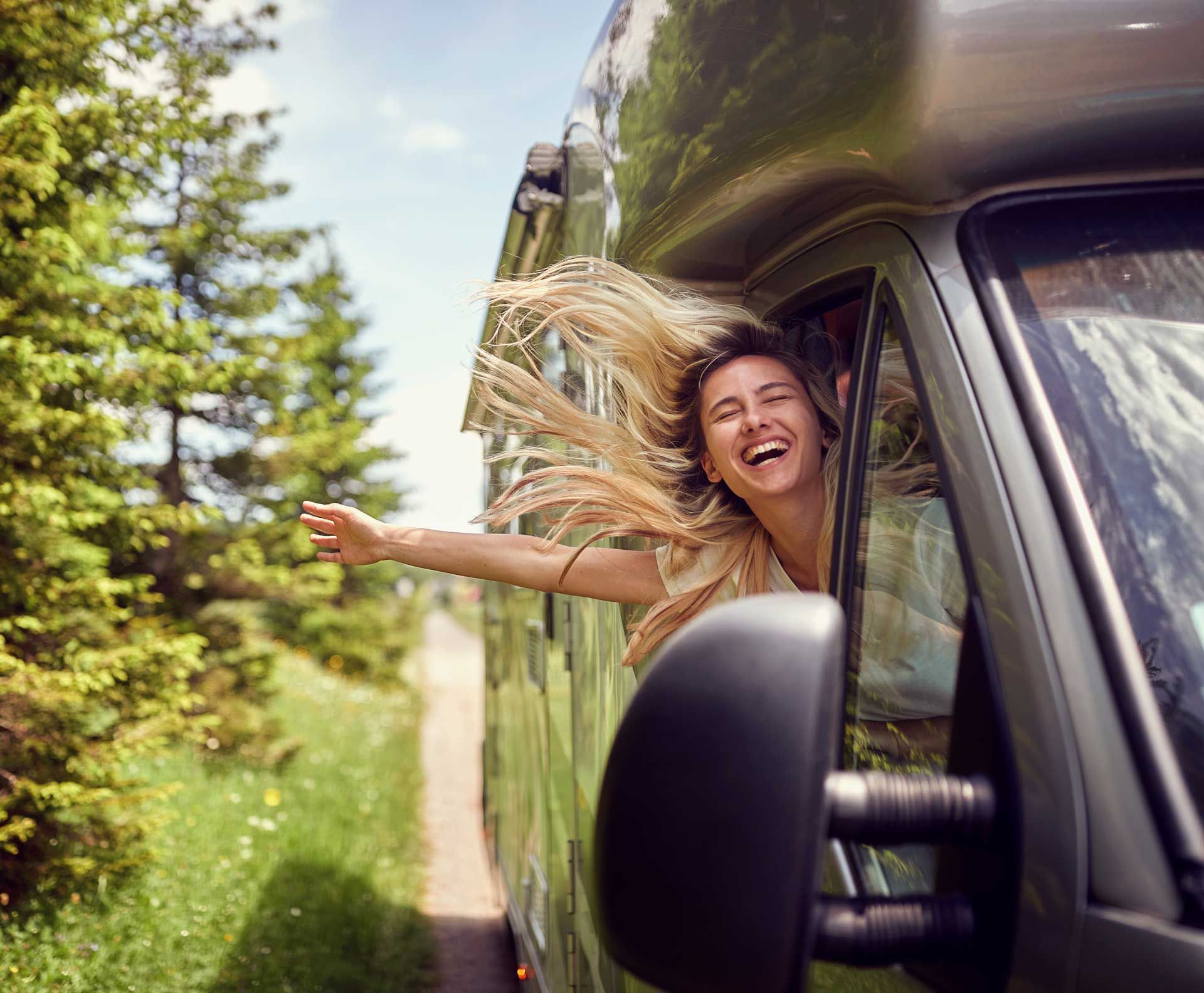 Woman with blonde hair and outstretched arm, smiles from the window of a moving camper van, scenic roadside view.