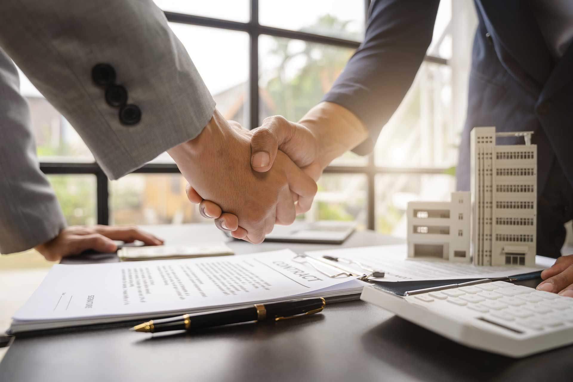 Two people shaking hands over a contract on a desk, with a model building and calculator nearby.