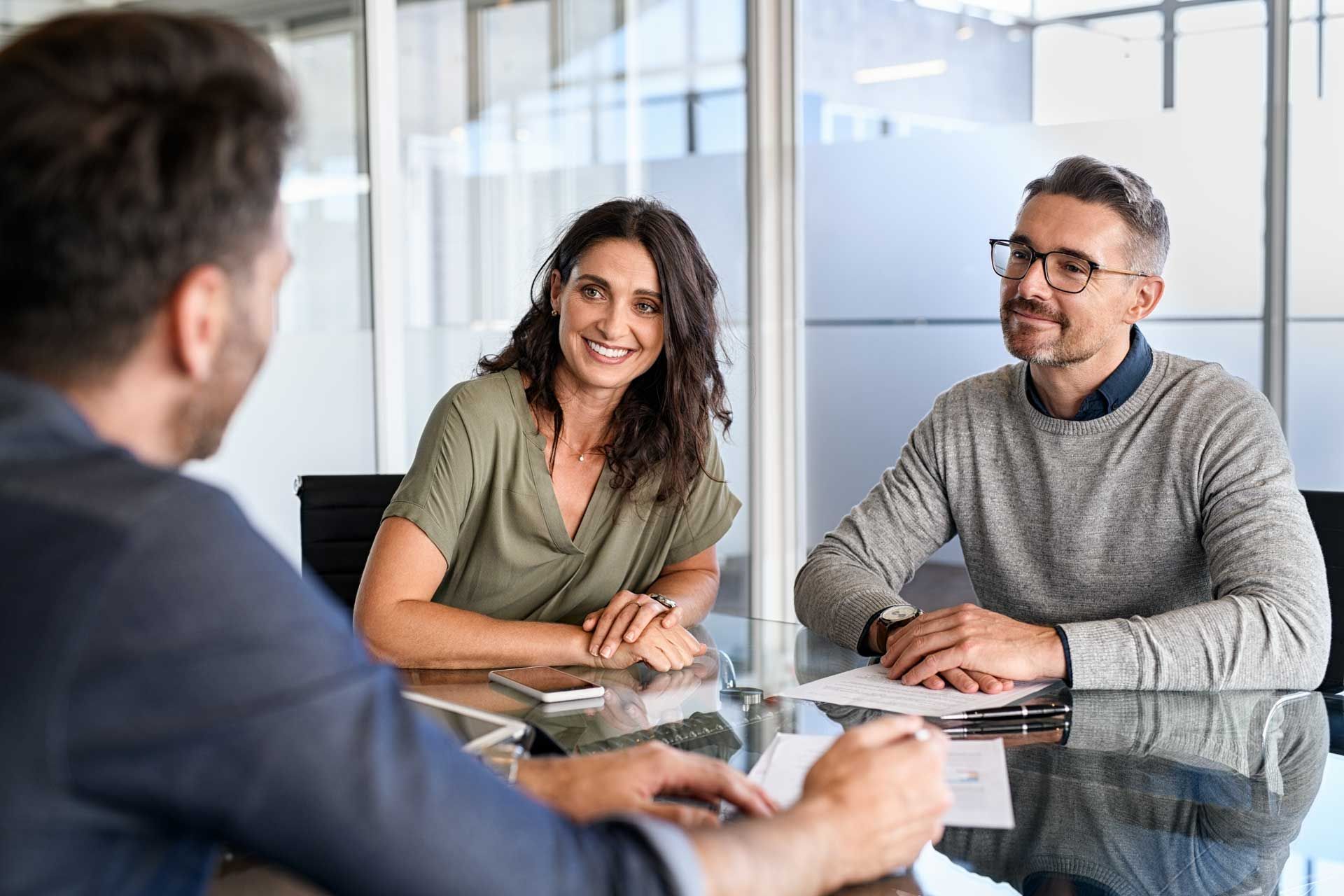 Two people smiling at person at a table in a glass-walled office. Documents on table. Two people smiling at person at a table in a glass-walled office. Documents on table.