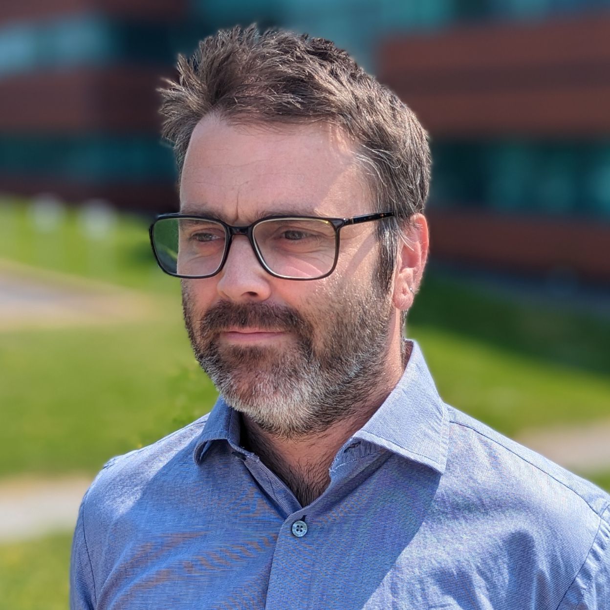 A man with a beard and glasses is standing in front of a building.