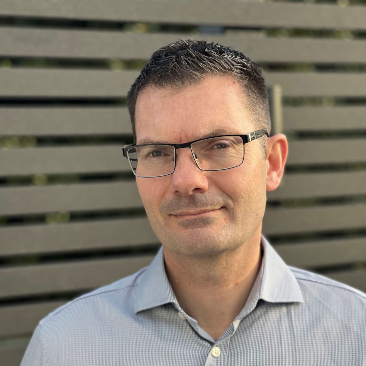 A man wearing glasses and a plaid shirt is standing in front of a wooden fence.