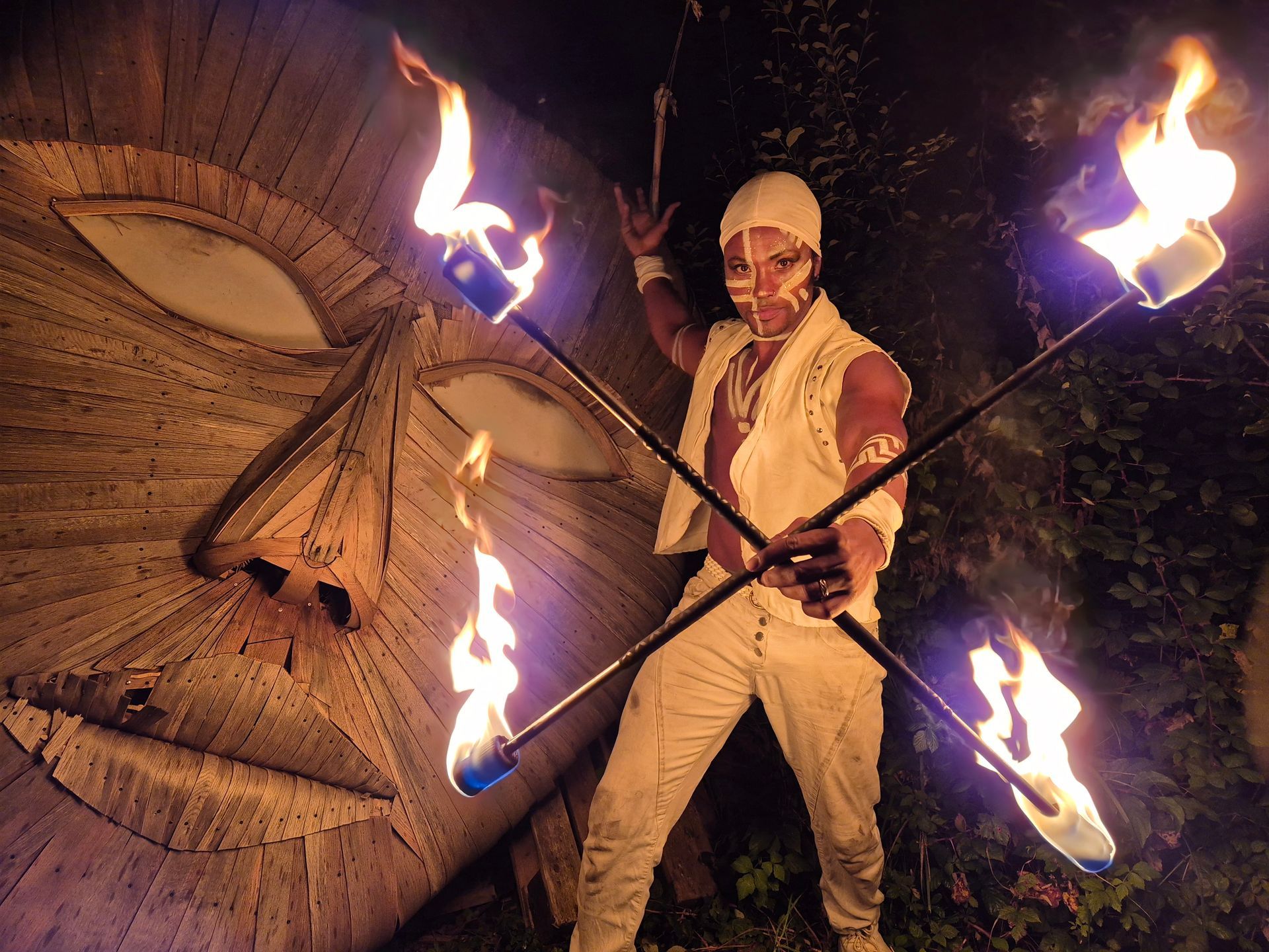Fire performer in white outfit holding flaming torches, standing before wooden mask.
