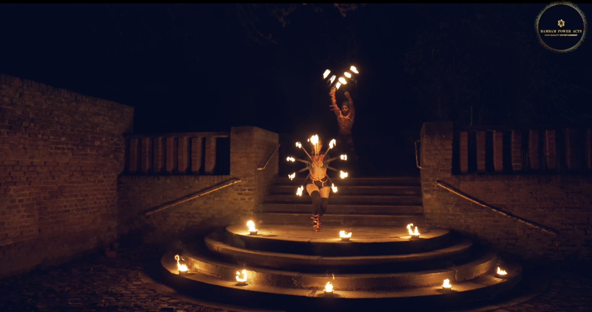 Fire dancer twirls fiery props on outdoor steps at night, surrounded by candles.