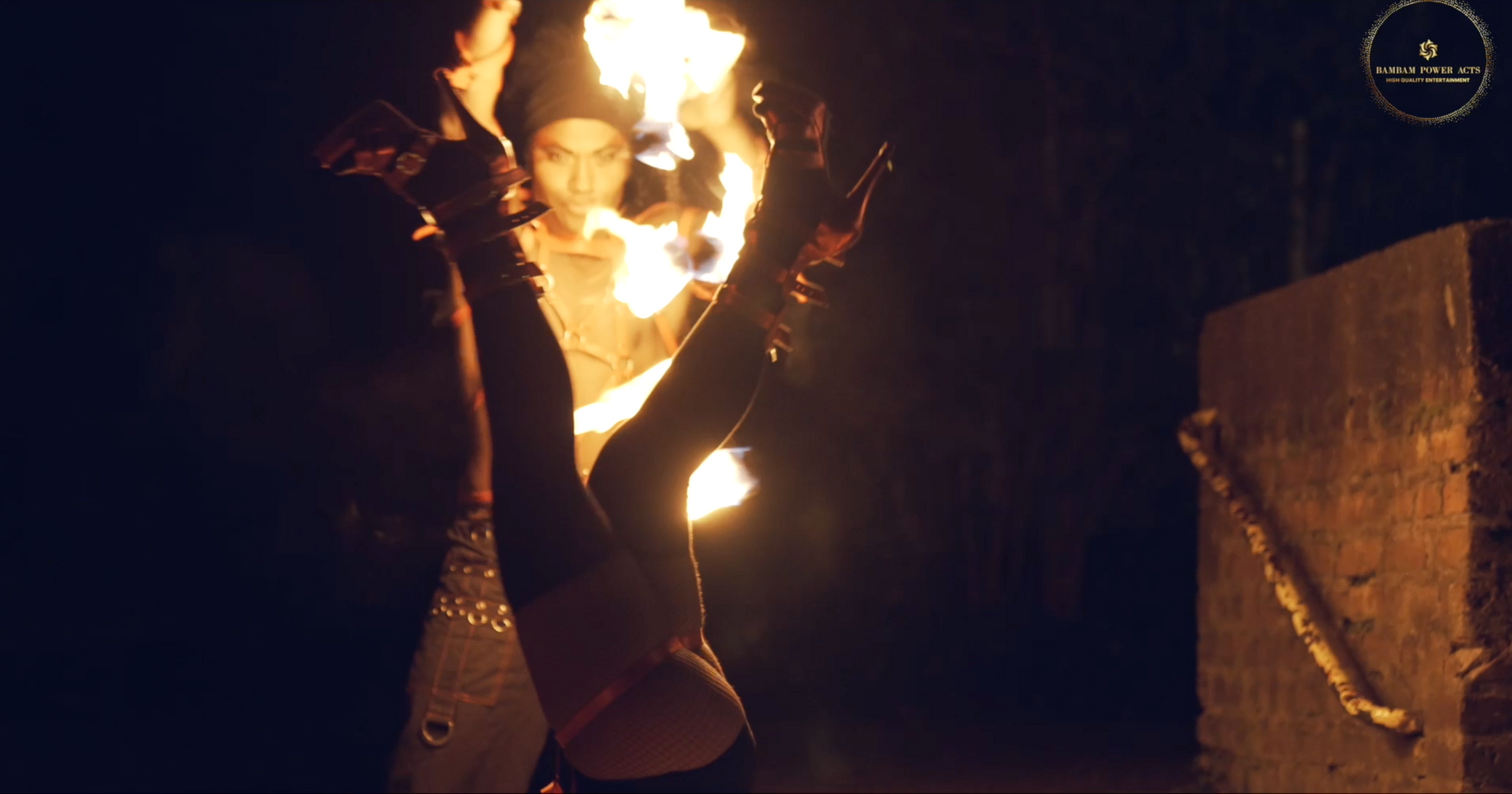 Person upside down, juggling fire. Another person watches, firelit setting.