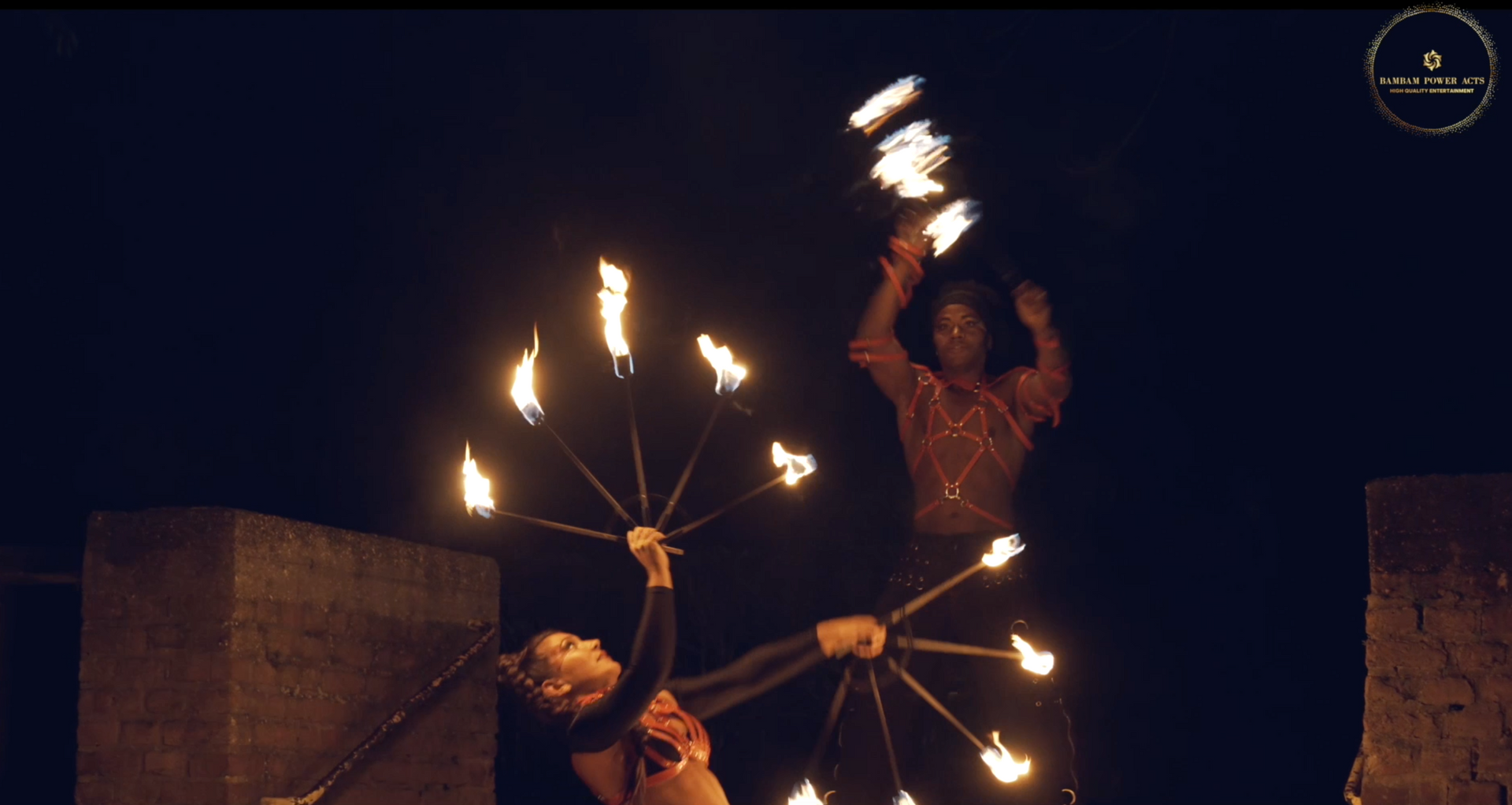 Two people performing fire dancing at night, illuminated by flames, against a dark background.