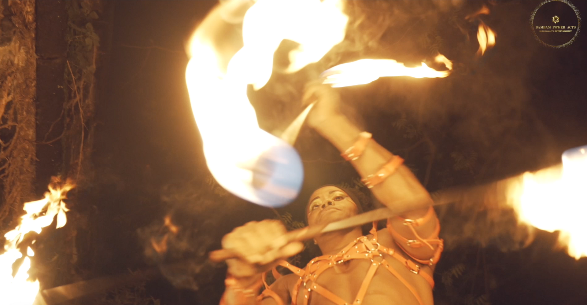 Fire performer twirling flaming batons in a dark cave, wearing orange attire, flames lighting the scene.
