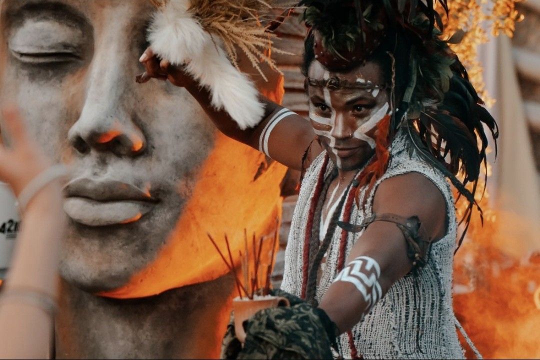 Man in tribal attire performing near a stone face sculpture, with red and white paint.