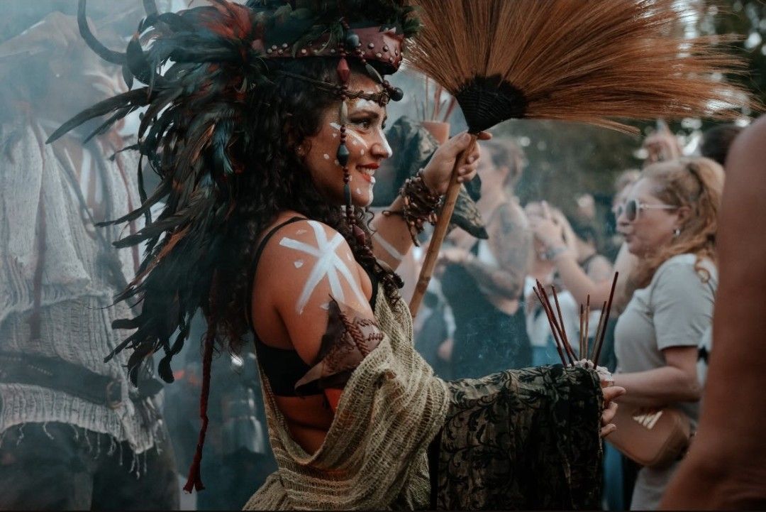 Woman in costume dancing, holding a broom and incense, at an outdoor gathering.