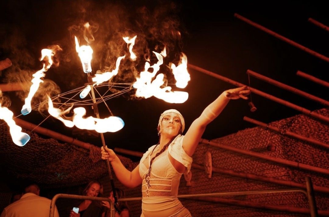 Woman in white outfit juggles flaming parasol at night.