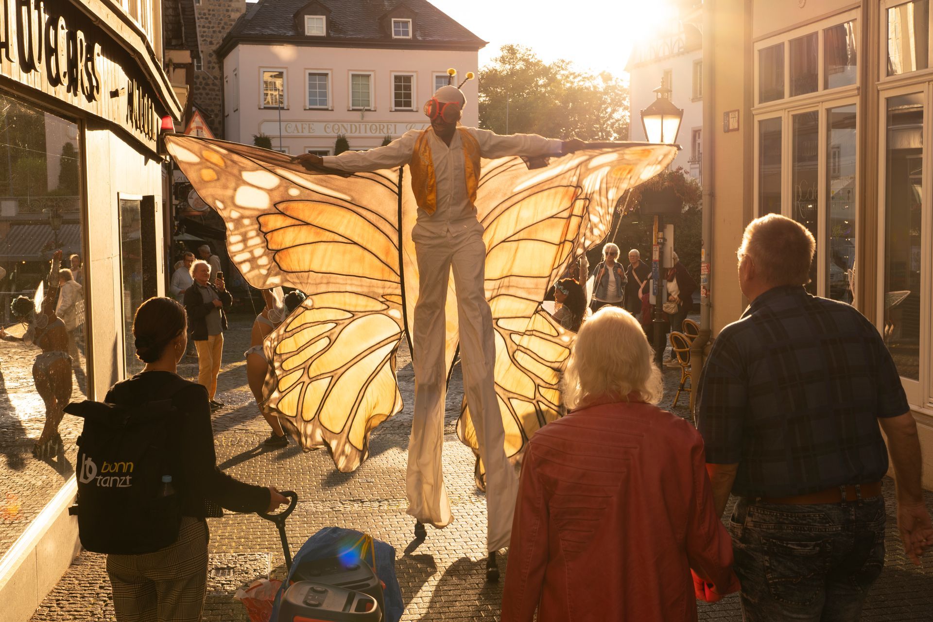 Person on stilts with butterfly wings, entertaining a crowd in a European street at sunset.