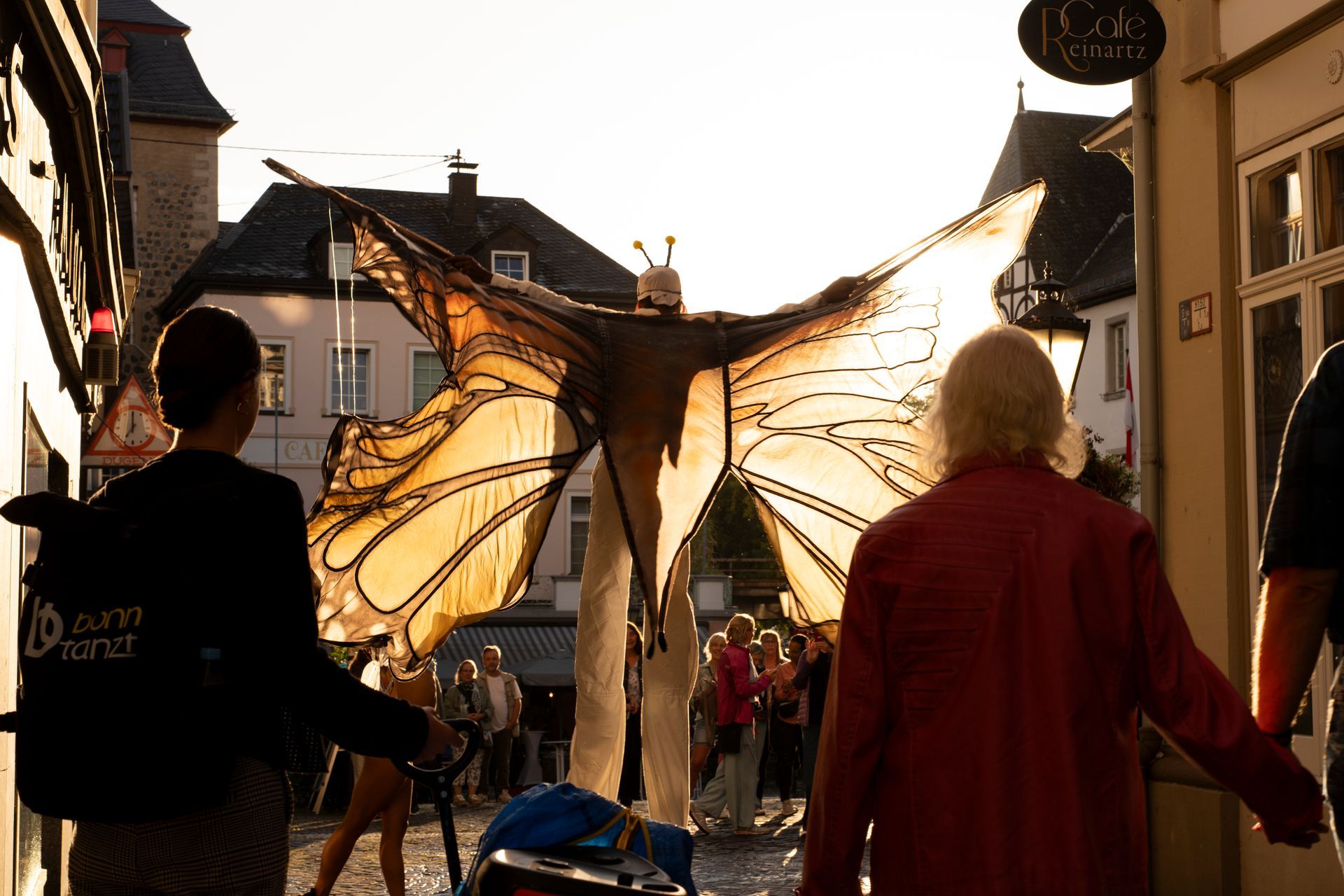 Giant butterfly performer in a street, illuminated by the sun, with people watching.