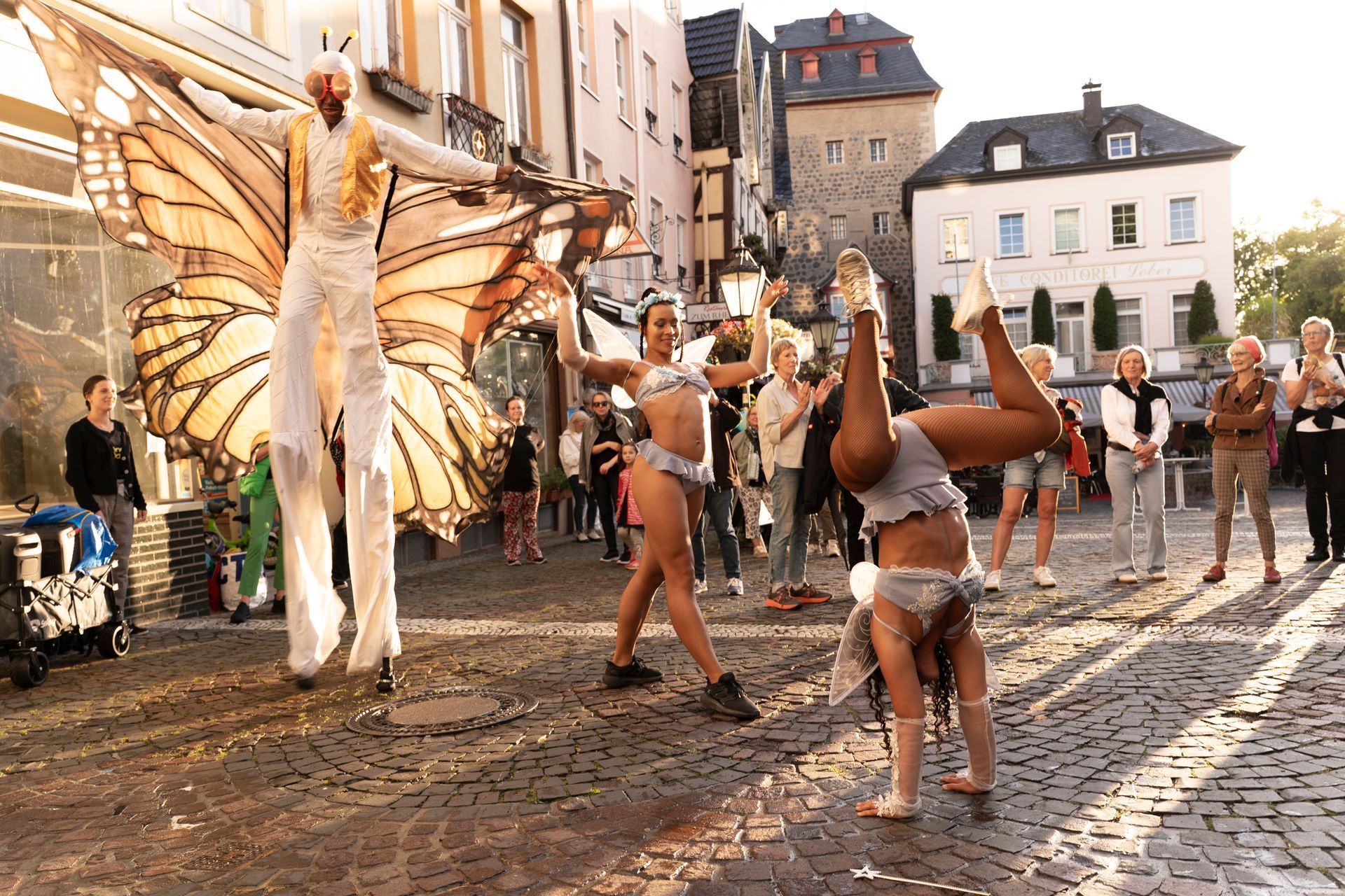 Street performers and crowd; a butterfly-winged stilt walker with two dancers; a woman doing a handstand.