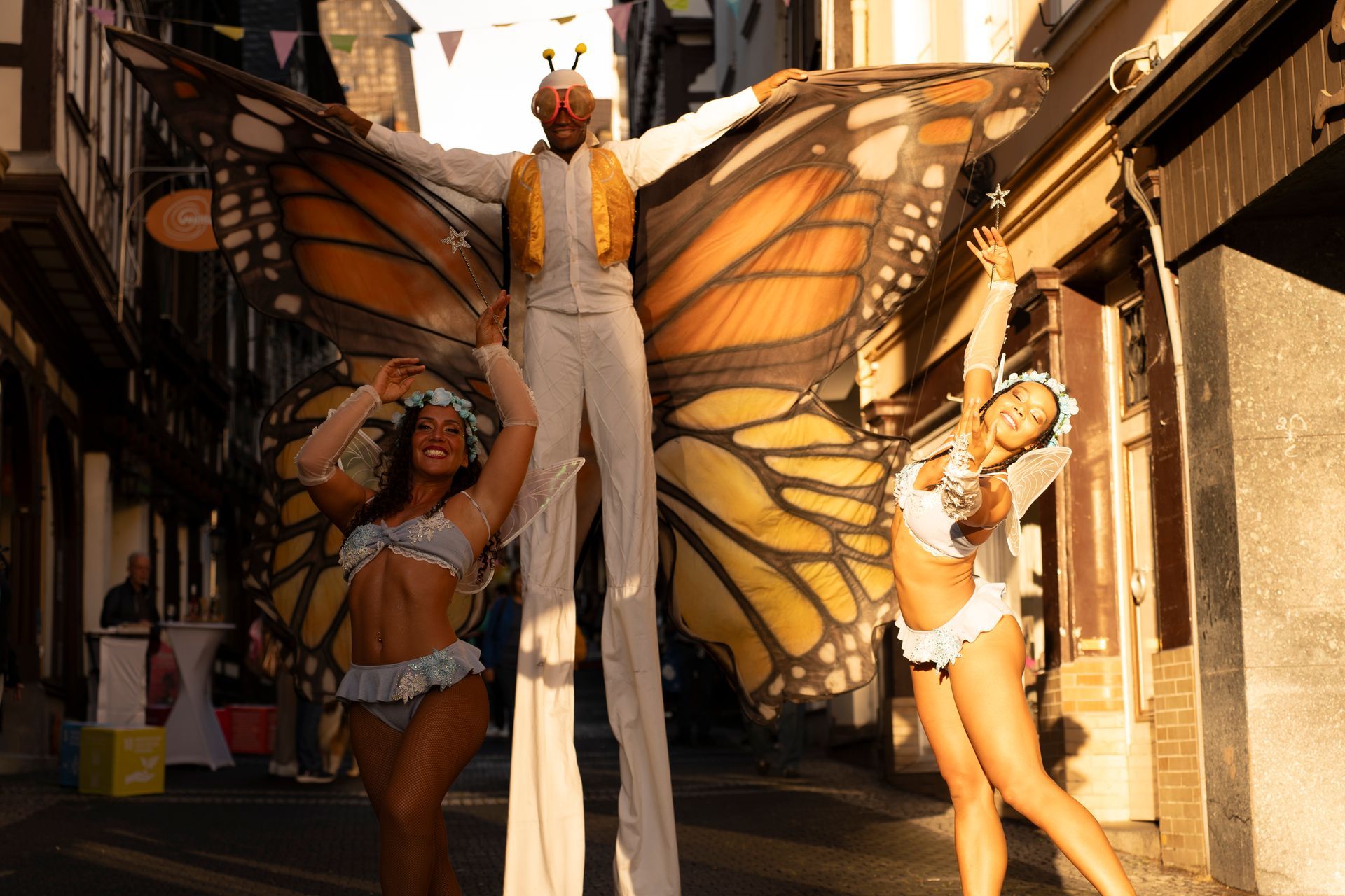 Person on stilts with butterfly wings flanked by two women in festive attire in a narrow street.
