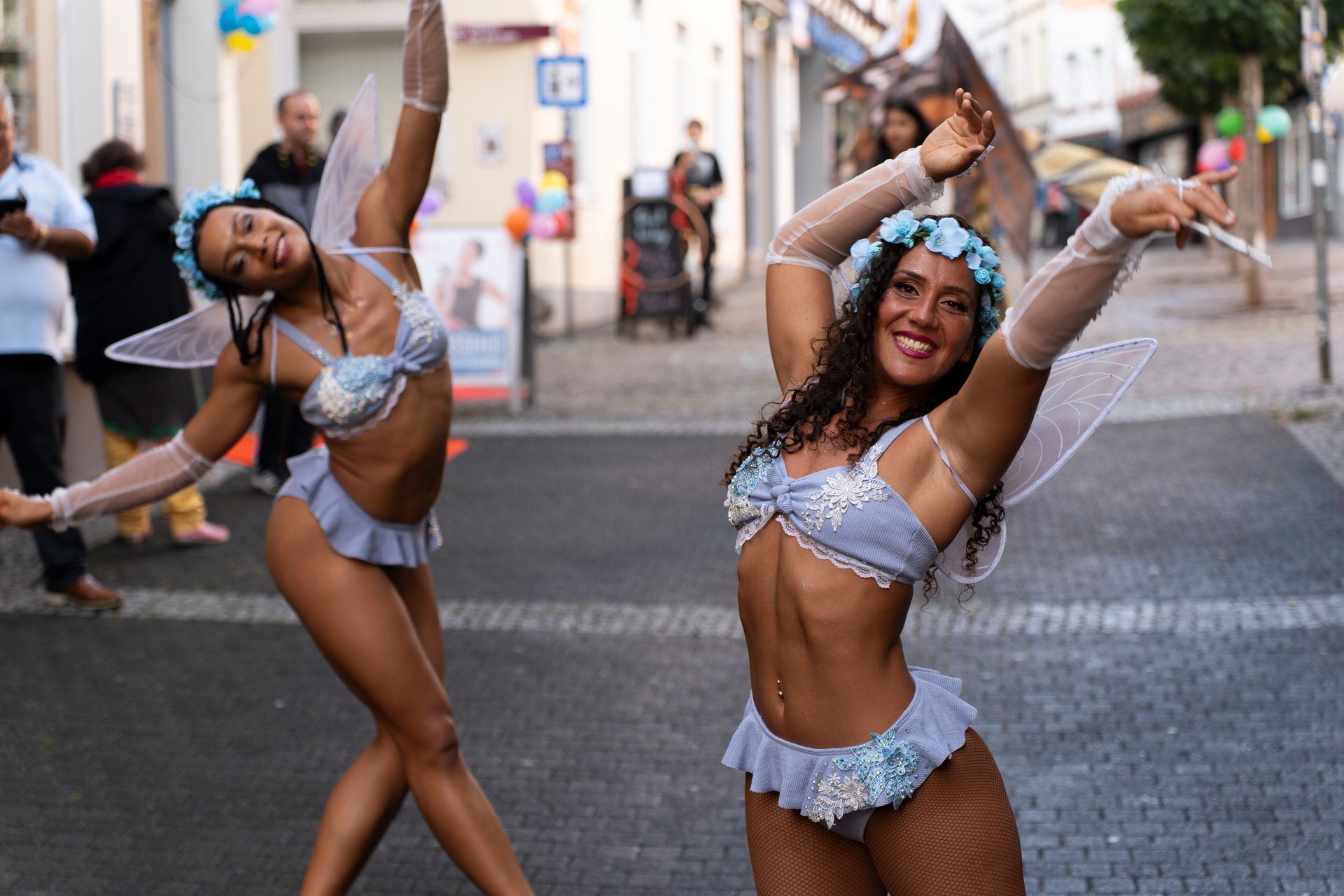 Two women in fairy costumes dance in a street, smiling and raising arms.