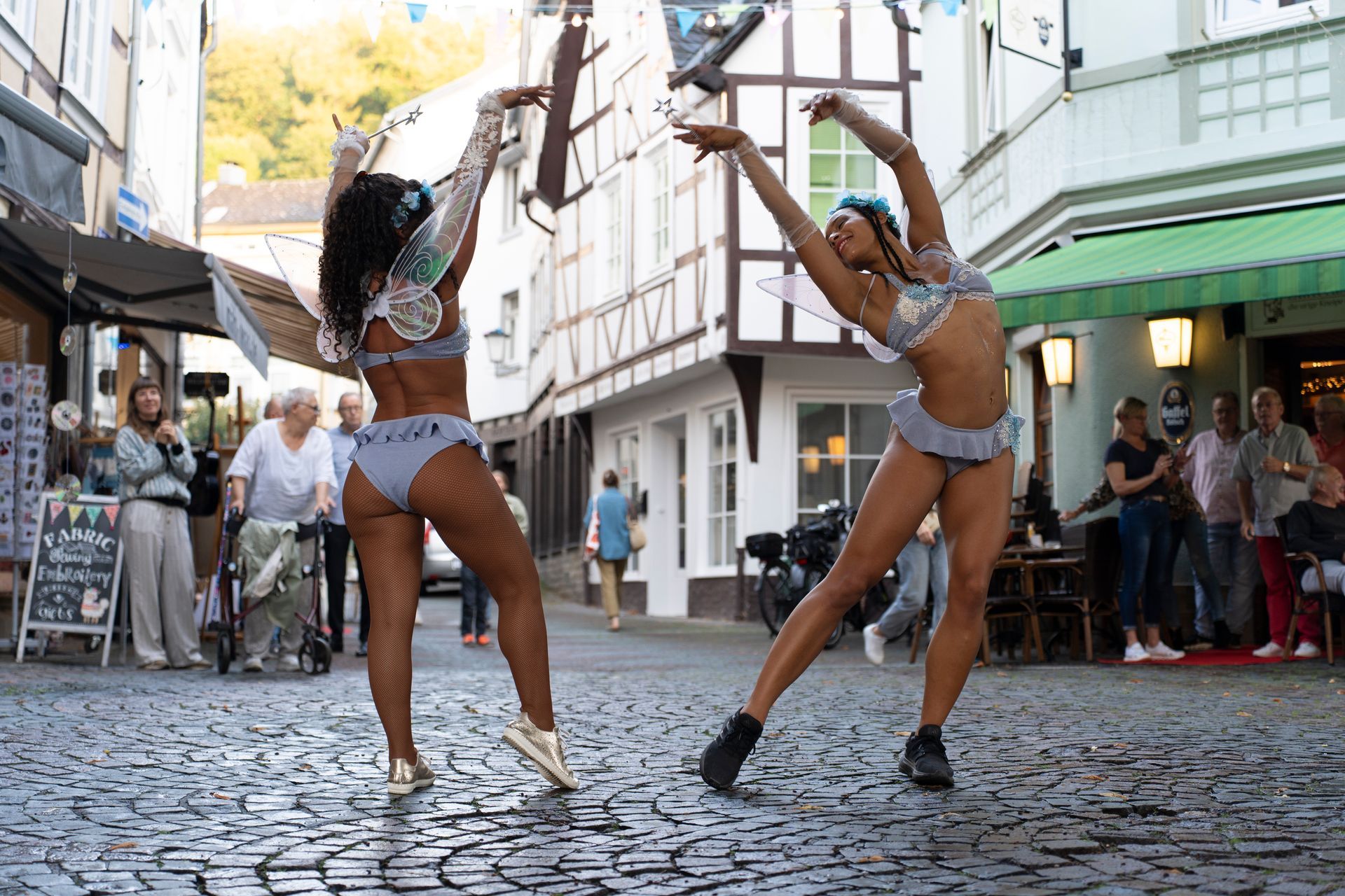 Two dancers in swimsuits perform on a cobblestone street. One wears gold shoes, the other black boots.
