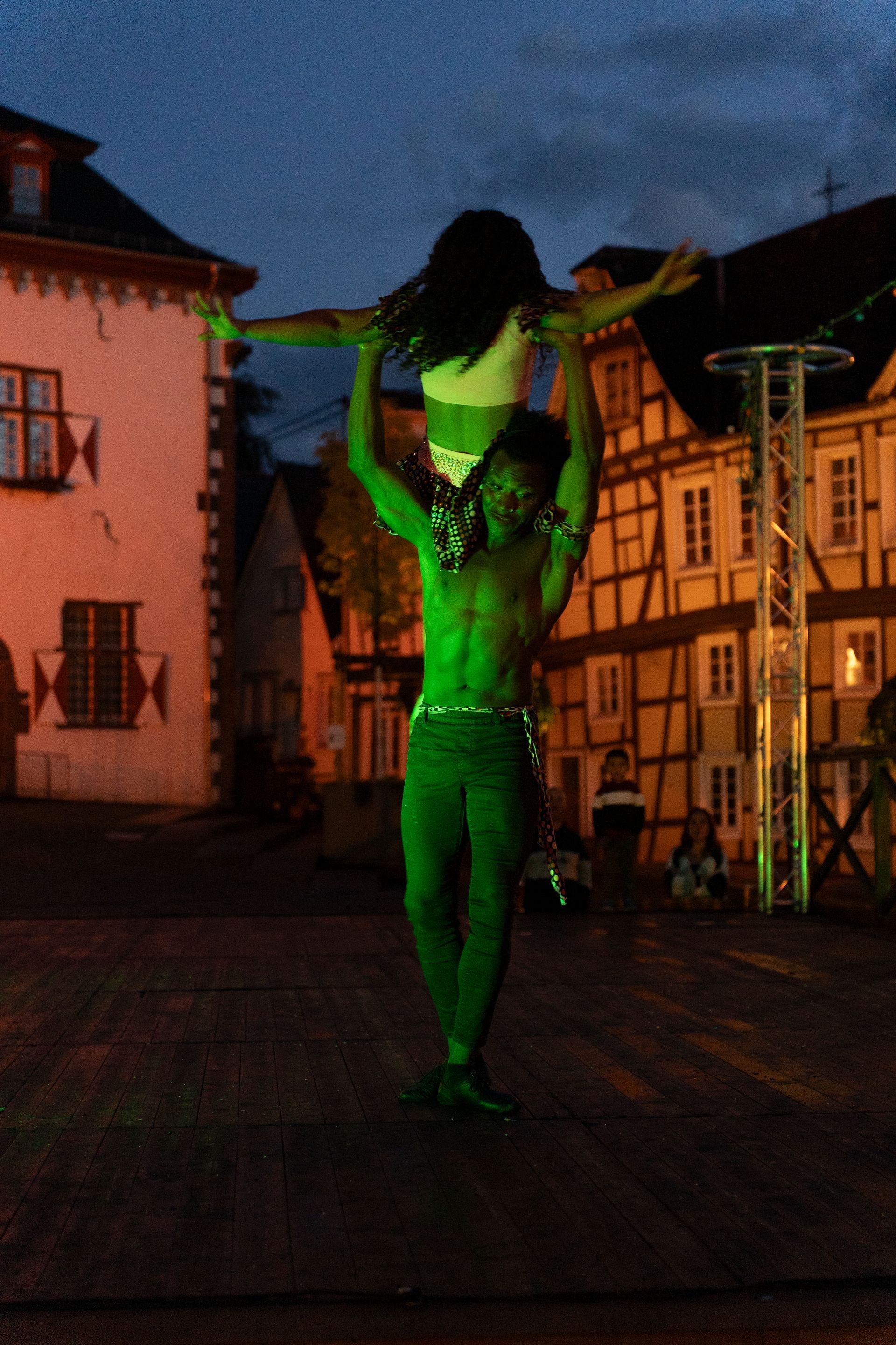 Person on stilts with butterfly wings flanked by two women in festive attire in a narrow street.