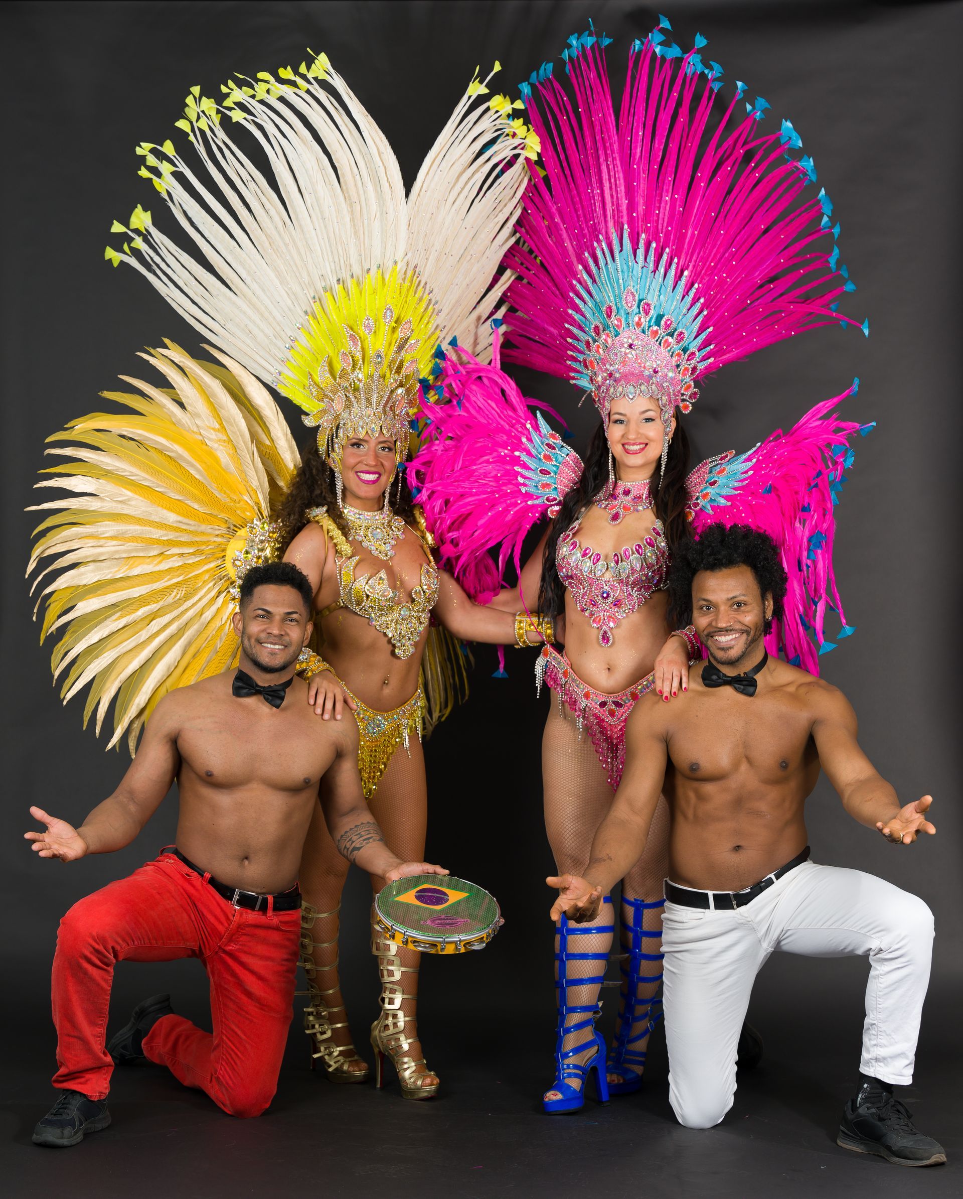 Two women in elaborate feathered costumes pose with two shirtless men, all smiling. Black background.