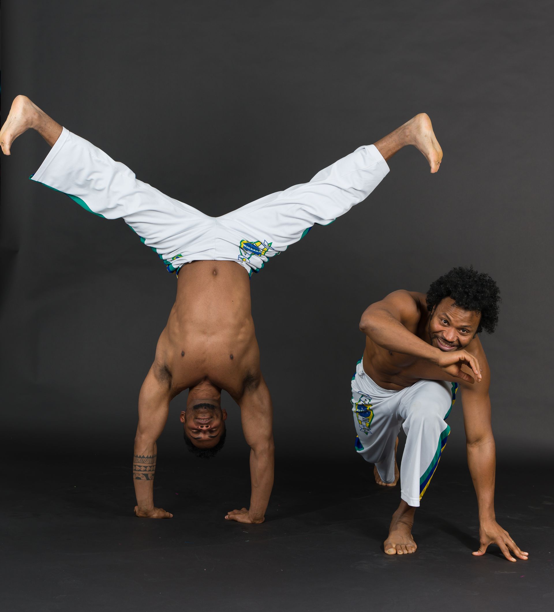 Two shirtless men doing Capoeira. One in a handstand, the other low to the ground, smiling, with white pants.