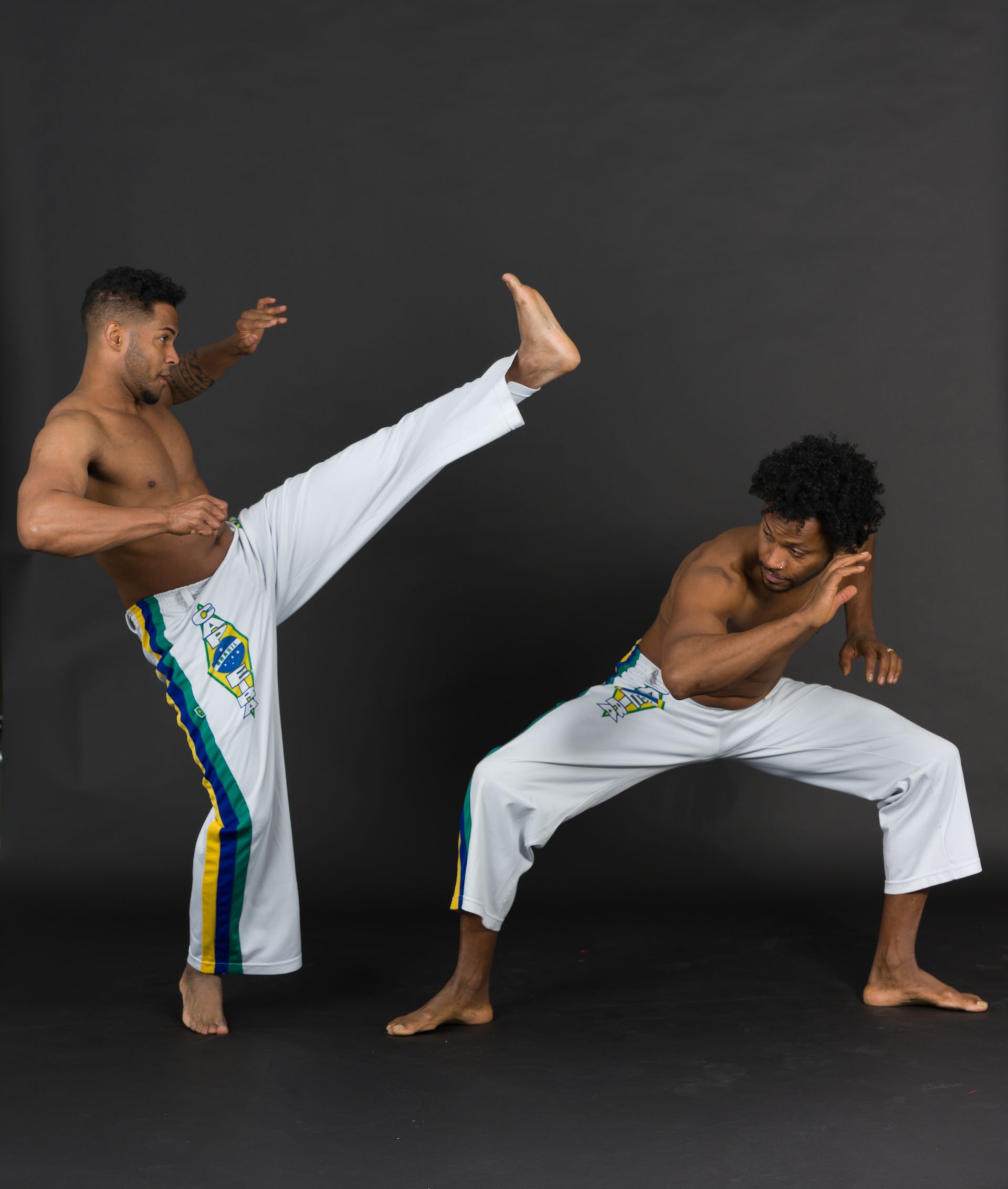 Two men in white pants practicing Capoeira kicks, one kicking high, the other low, in front of a dark background.