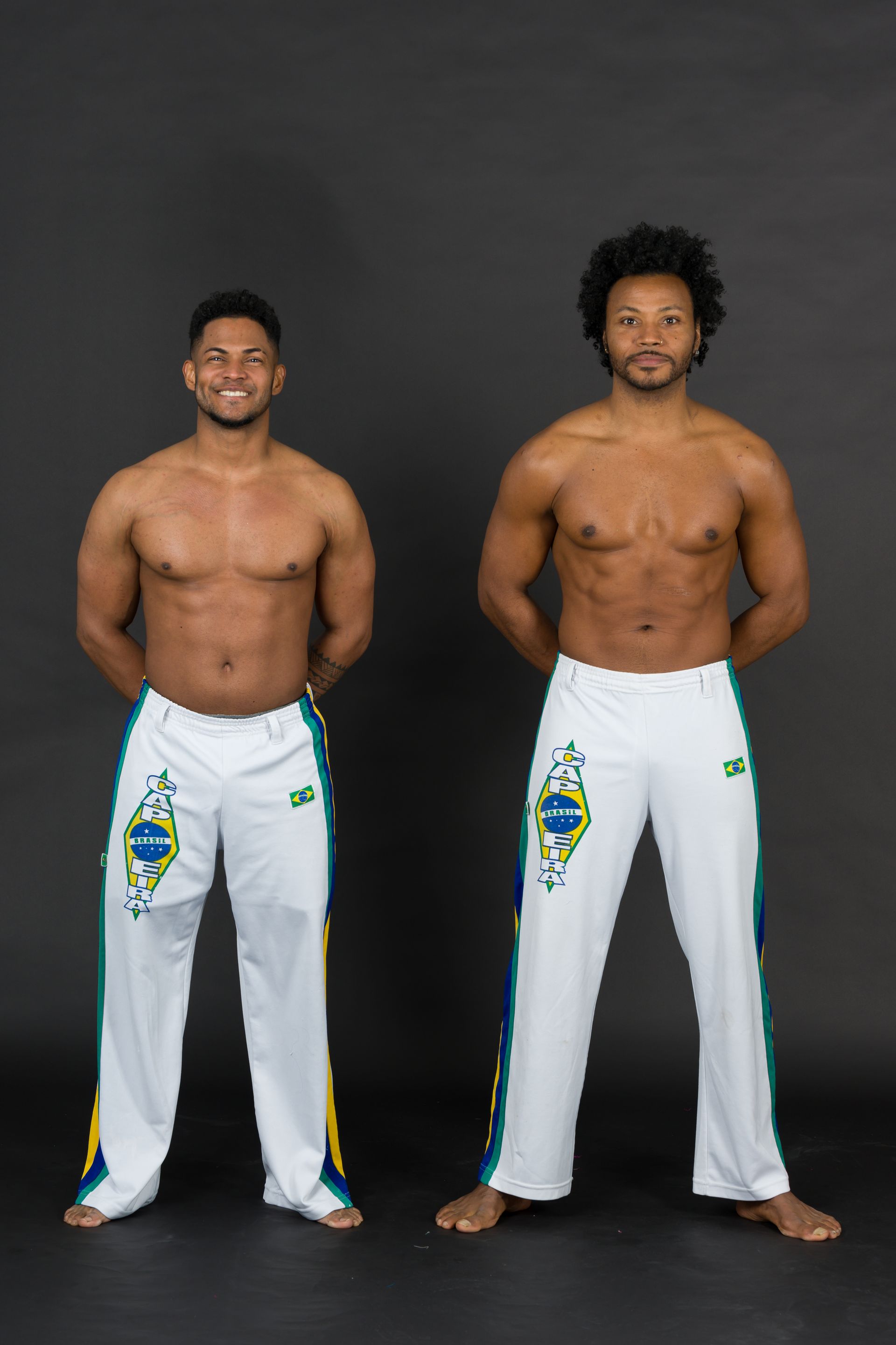 Two shirtless men in white capoeira pants stand, hands behind their backs, on a gray backdrop.