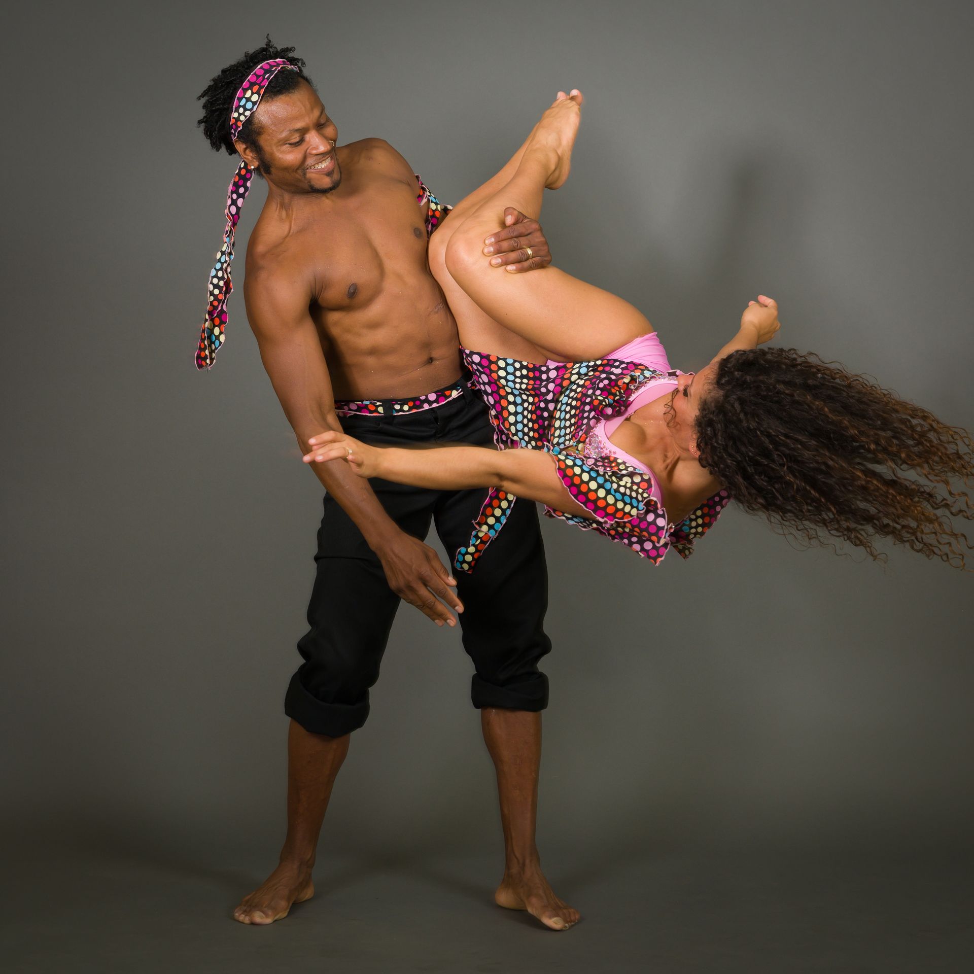 Man holding a woman, both smiling, performing a dance move. Gray background.