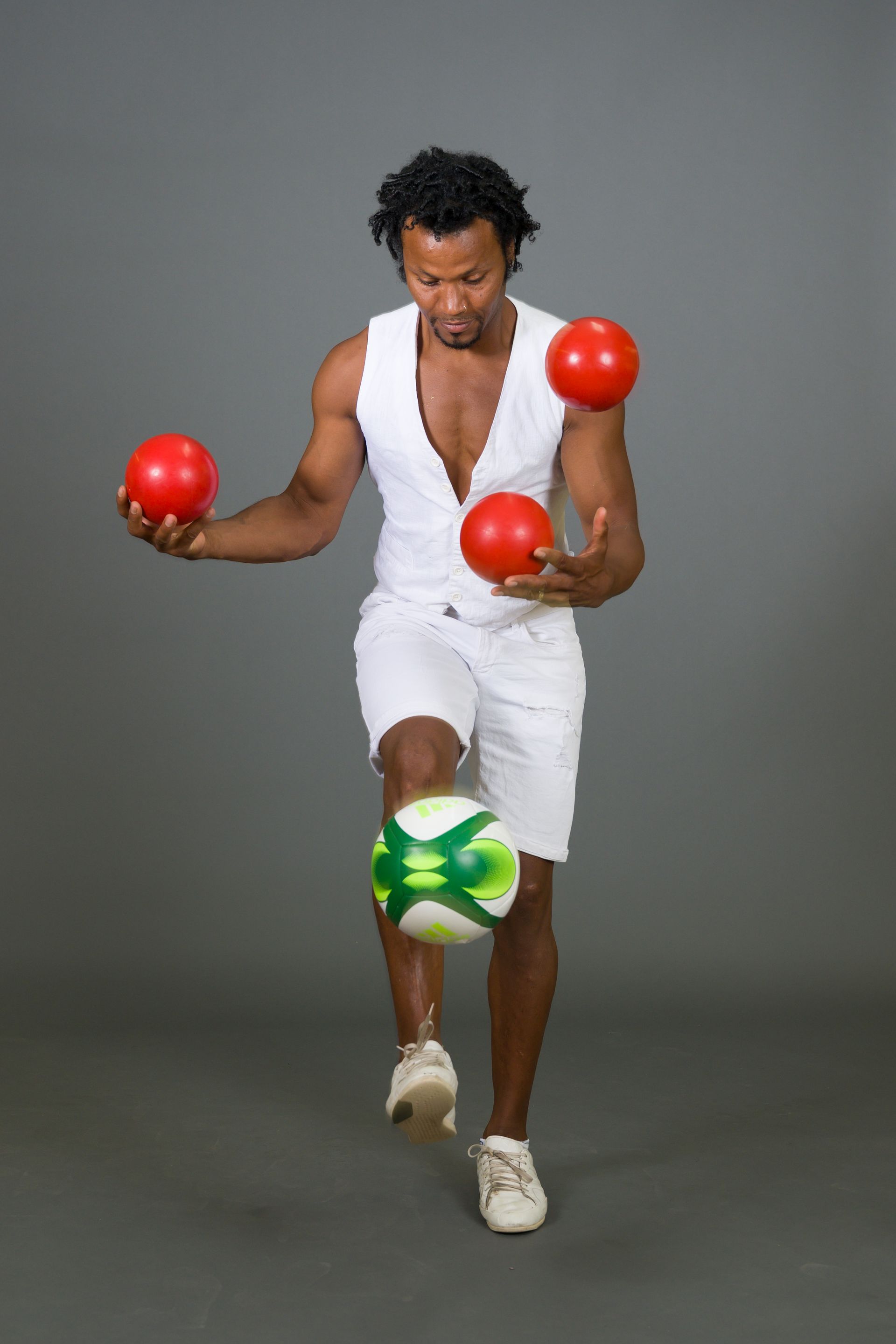 Man juggling three red balls and soccer ball. Wearing white outfit, against gray background.