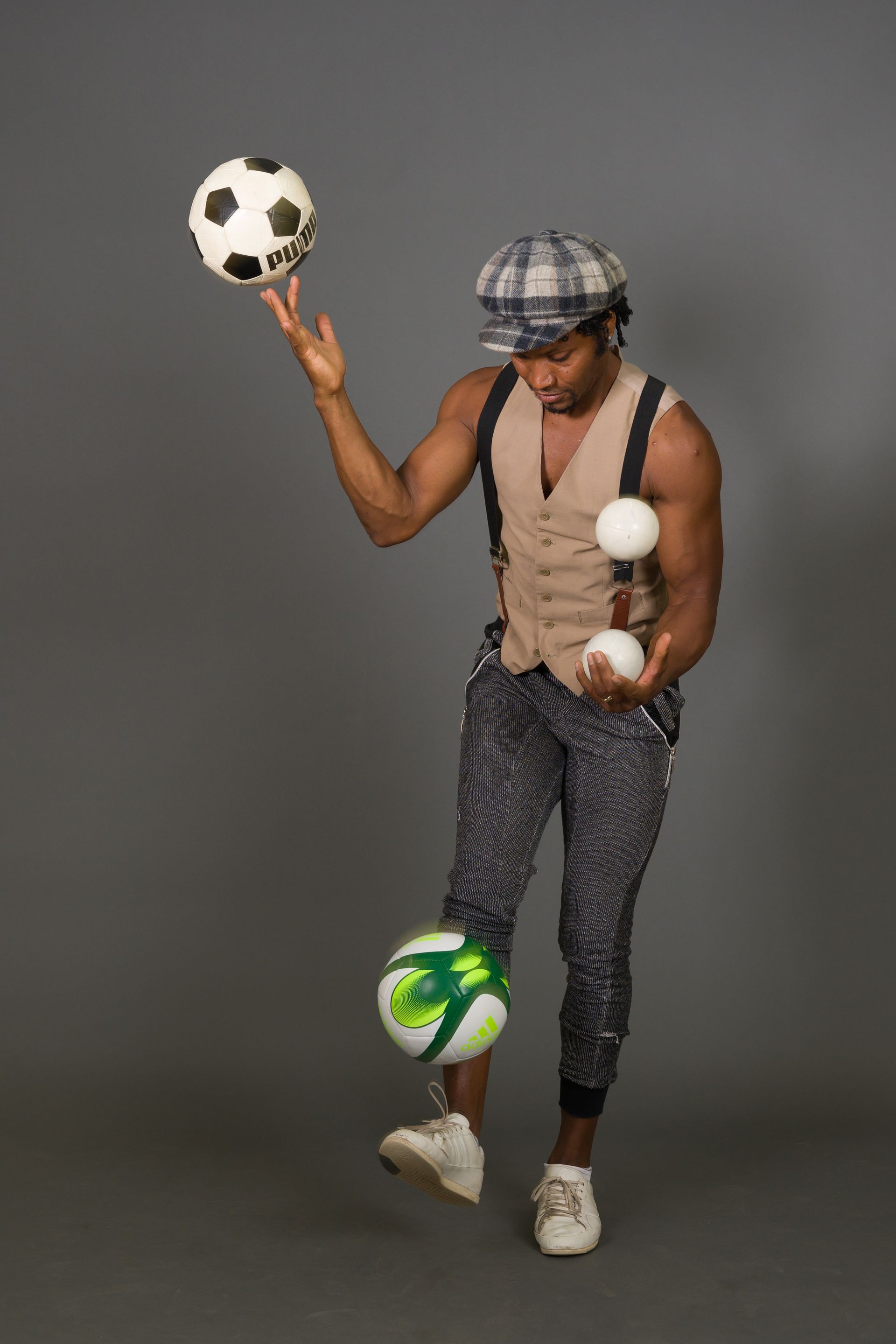 Man juggling soccer balls and white balls, wearing a cap and suspenders.