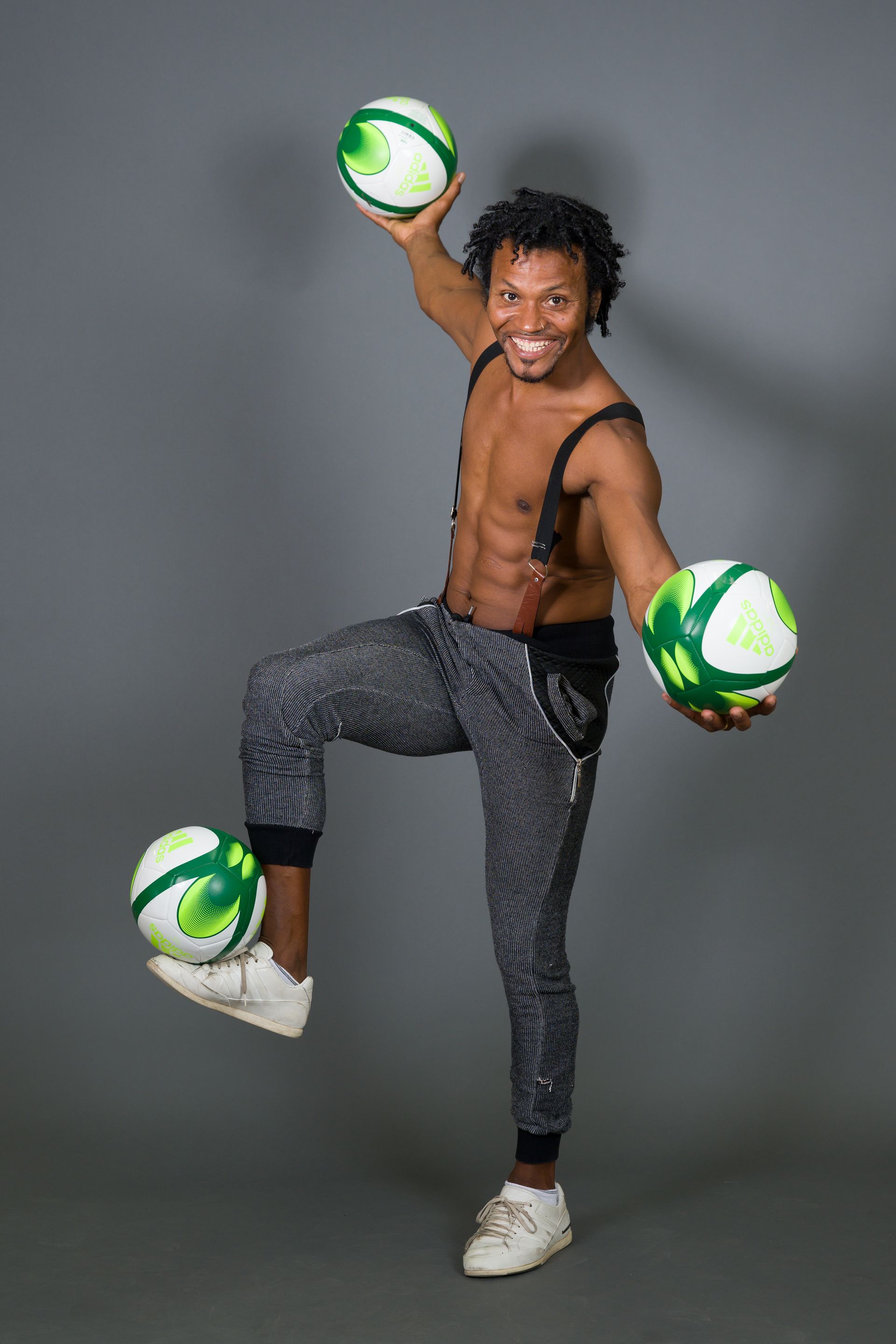 Man juggling three balls with one on his foot, smiling, wearing suspenders, black pants, and white shoes.