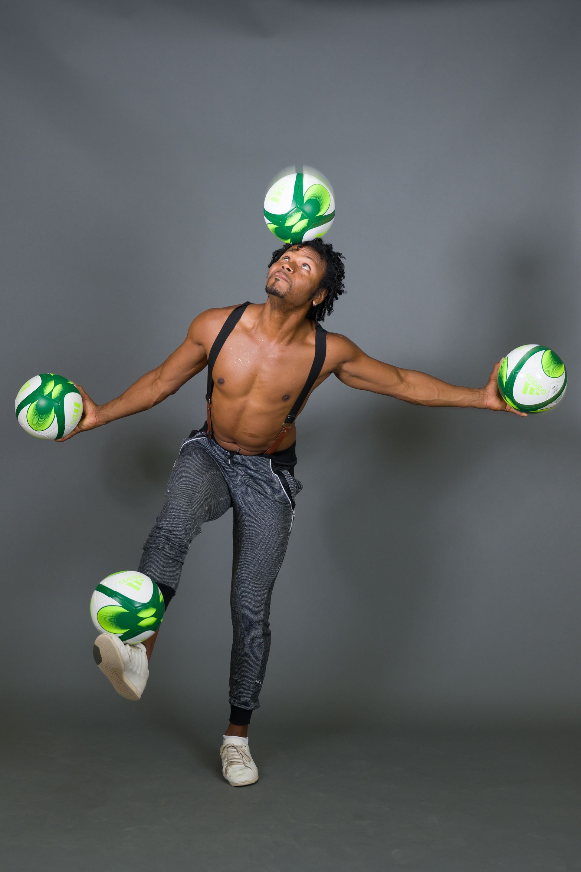 Man juggling five soccer balls, balanced on foot and head, grey background.