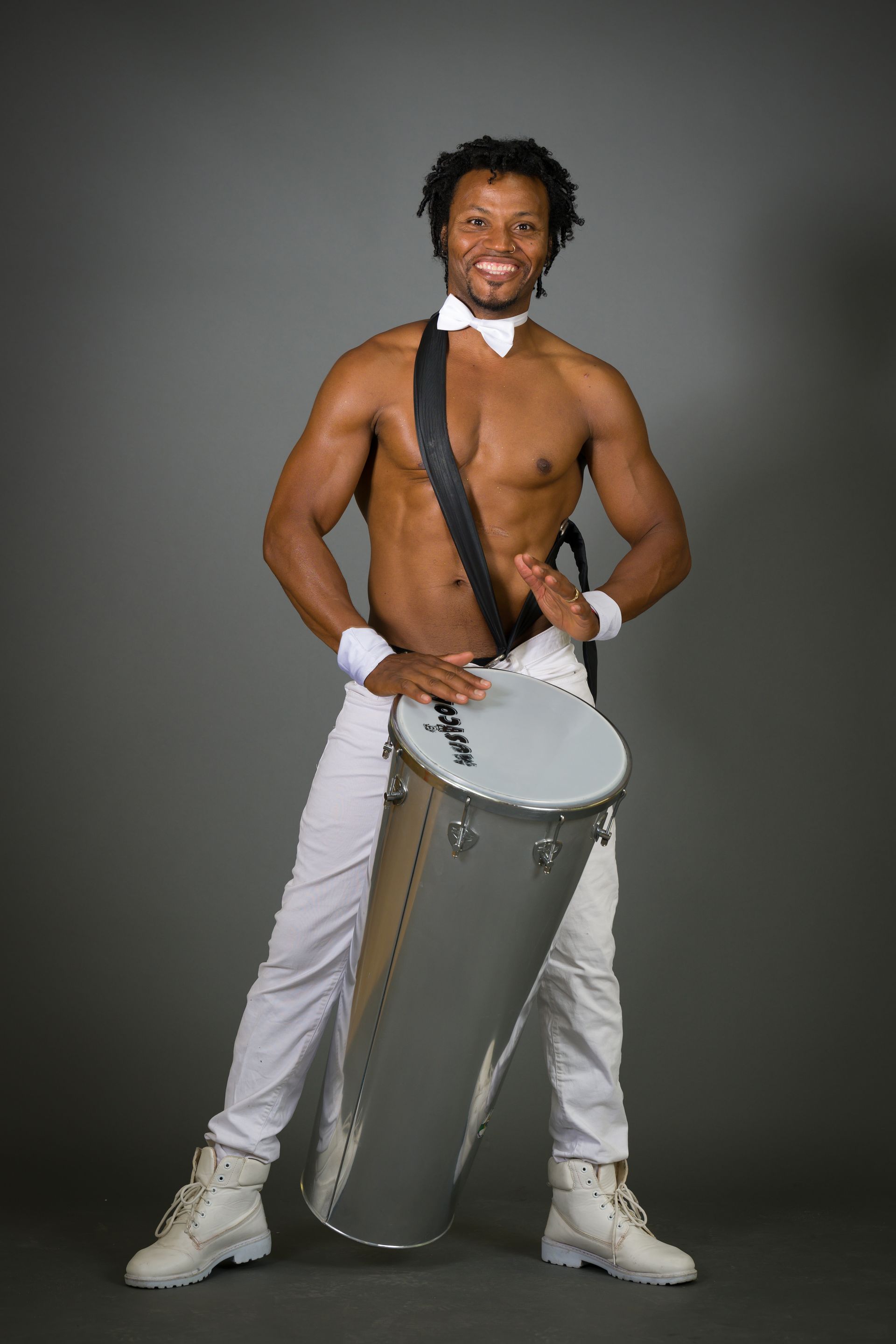 Man playing a drum, shirtless, in white pants and shoes, smiling.