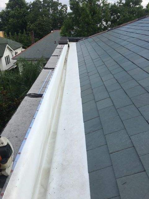 Gray shingled roof with two chimneys against a blue sky and partially visible building exterior.
