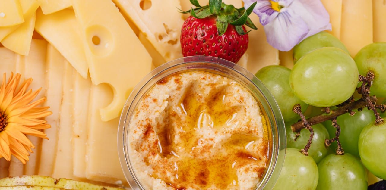 Una tabla de quesos y frutas con hummus, uvas, lonchas de queso y una fresa.