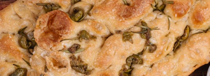 Primer plano de una focaccia cubierta con aceitunas verdes y hierbas. La corteza está dorada.
