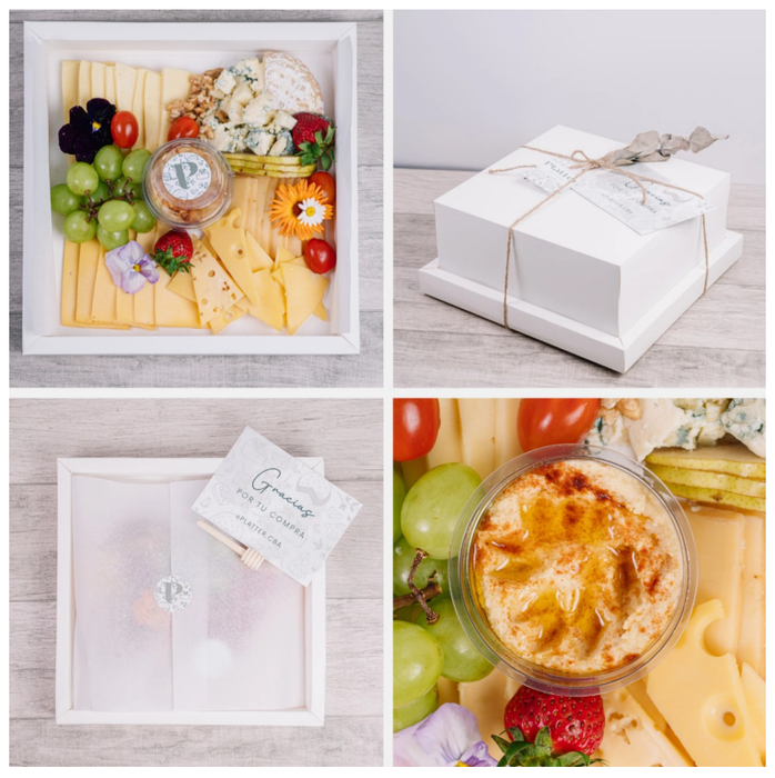Tabla de embutidos en caja blanca; incluye queso, uvas, hummus, flores y tomates.