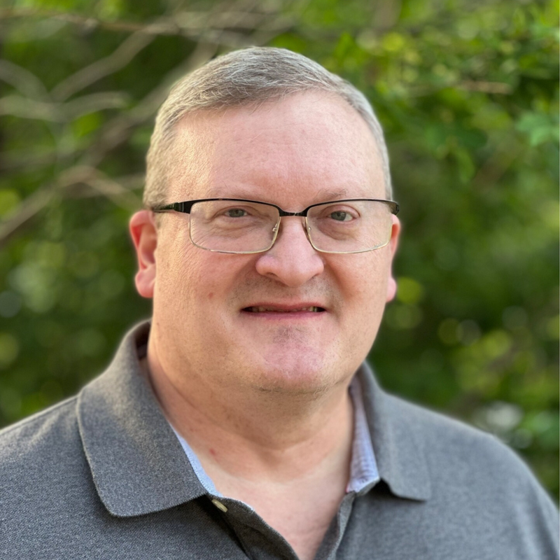 Man wearing glasses and a gray collared shirt smiles outdoors.