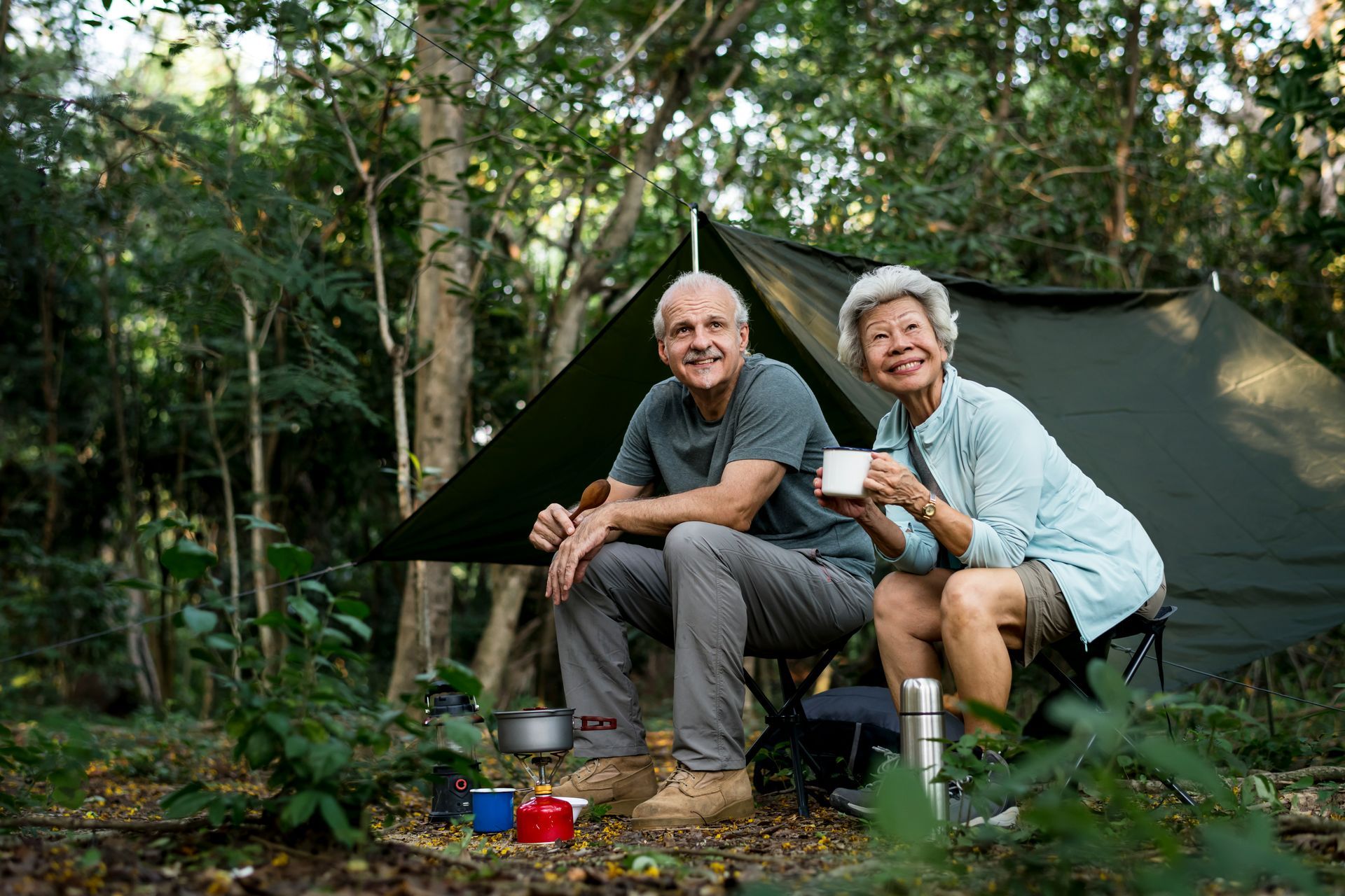 Couple sitting by a tent in a forest, smiling. Man in grey, woman in blue holding a mug.