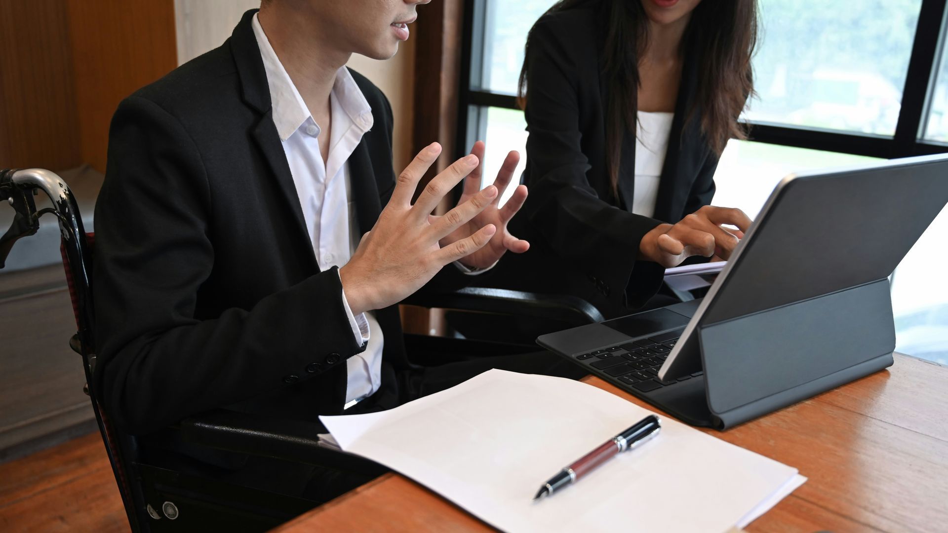 Two people in business attire at a desk, looking at a laptop and documents. One gestures with hands.