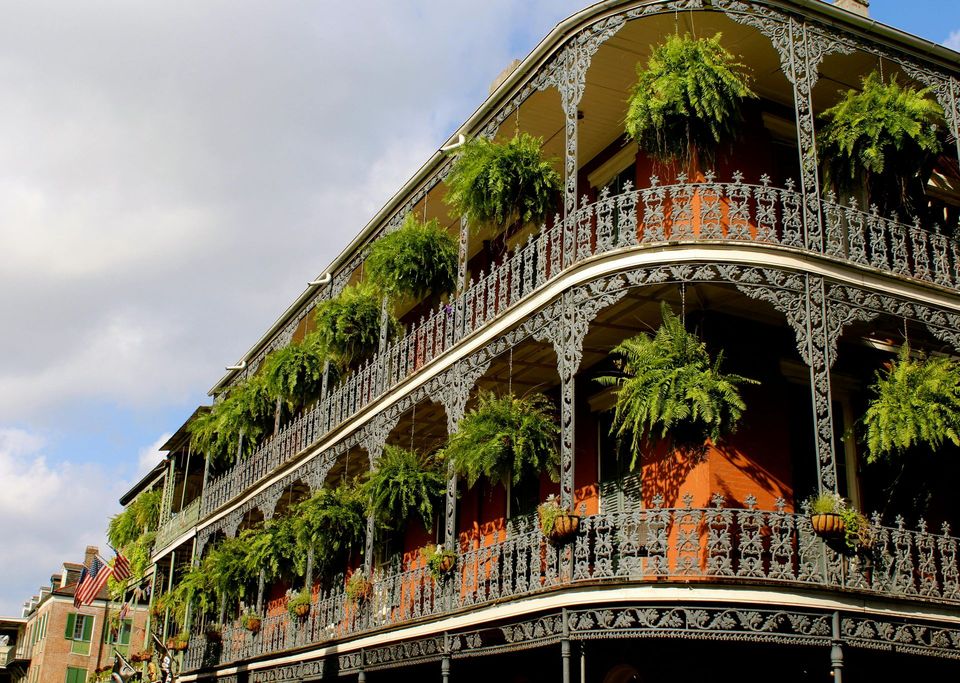 Ornate wrought-iron balconies of a New Orleans building, adorned with hanging ferns. Blue sky with clouds.