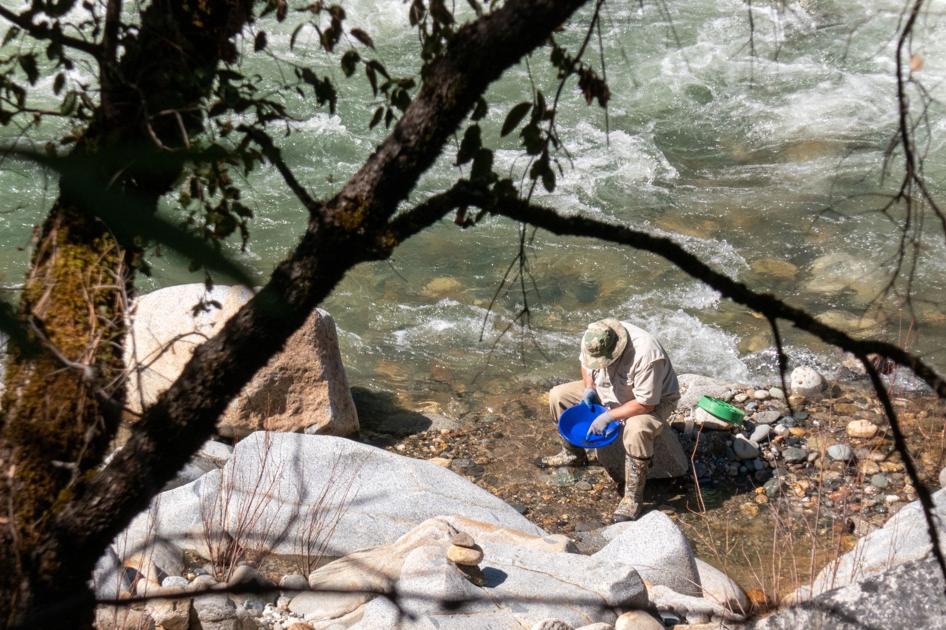 Man panning for gold in a river, using a blue pan and green sluice box. Sunlight on the water.