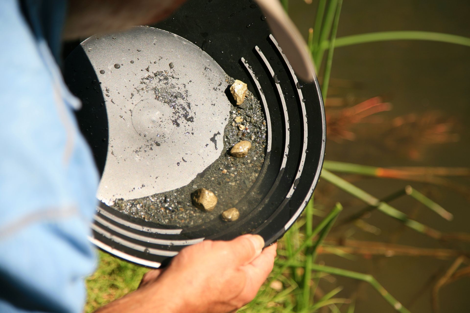 Person panning for gold in a black pan near a body of water.