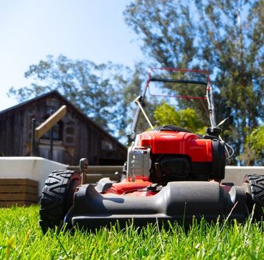 A bright red lawnmower sits on a vibrant green lawn in front of a rustic wooden house under a clear blue sky.