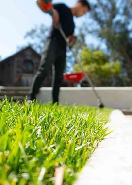 A blurred person in black clothes trims grass along a concrete edge with a string trimmer.