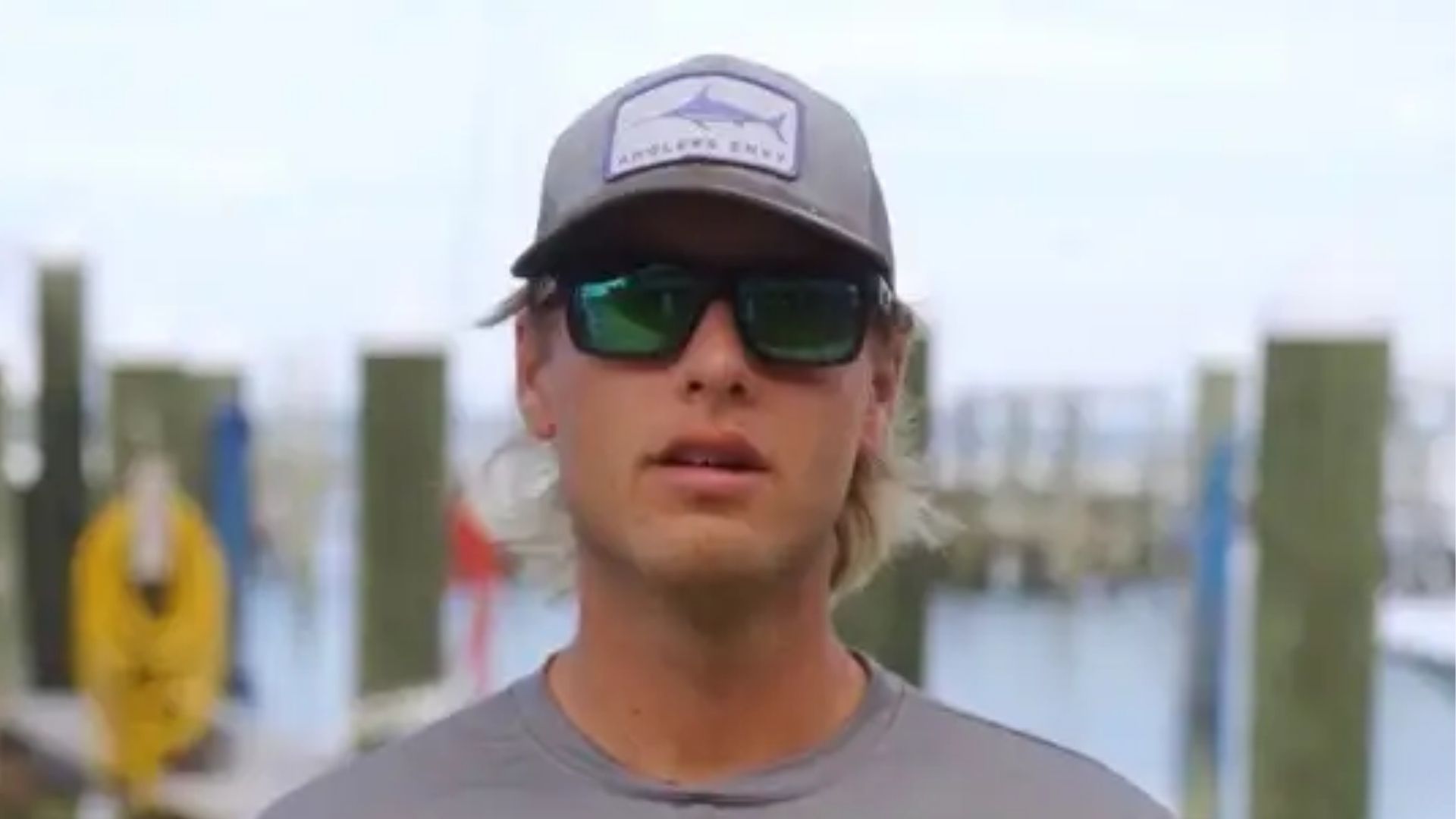 A person wearing a grey cap and sunglasses stands in front of wooden dock pilings at a marina.