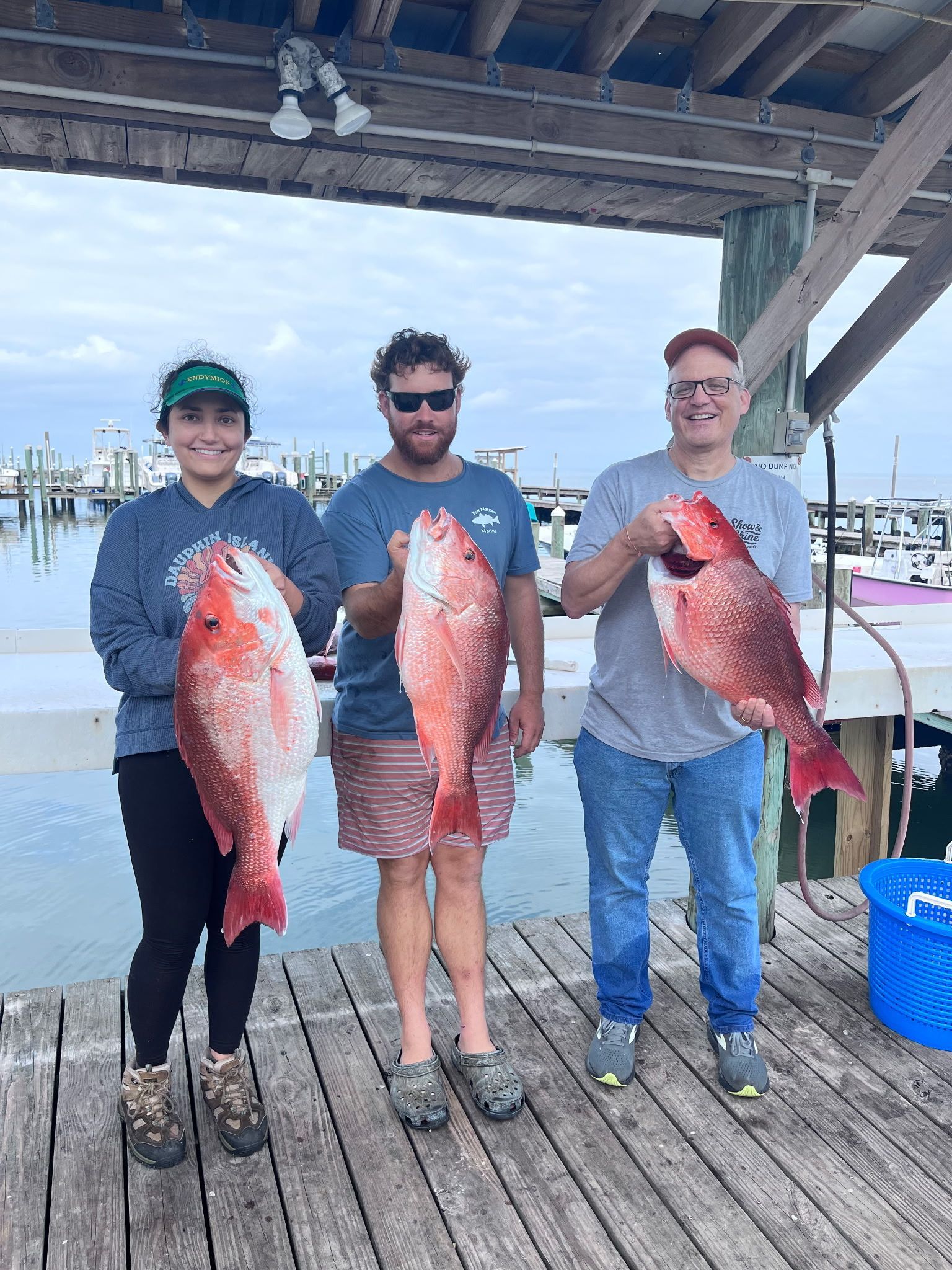 Three people on a dock holding large red fish, with boats and water in the background.